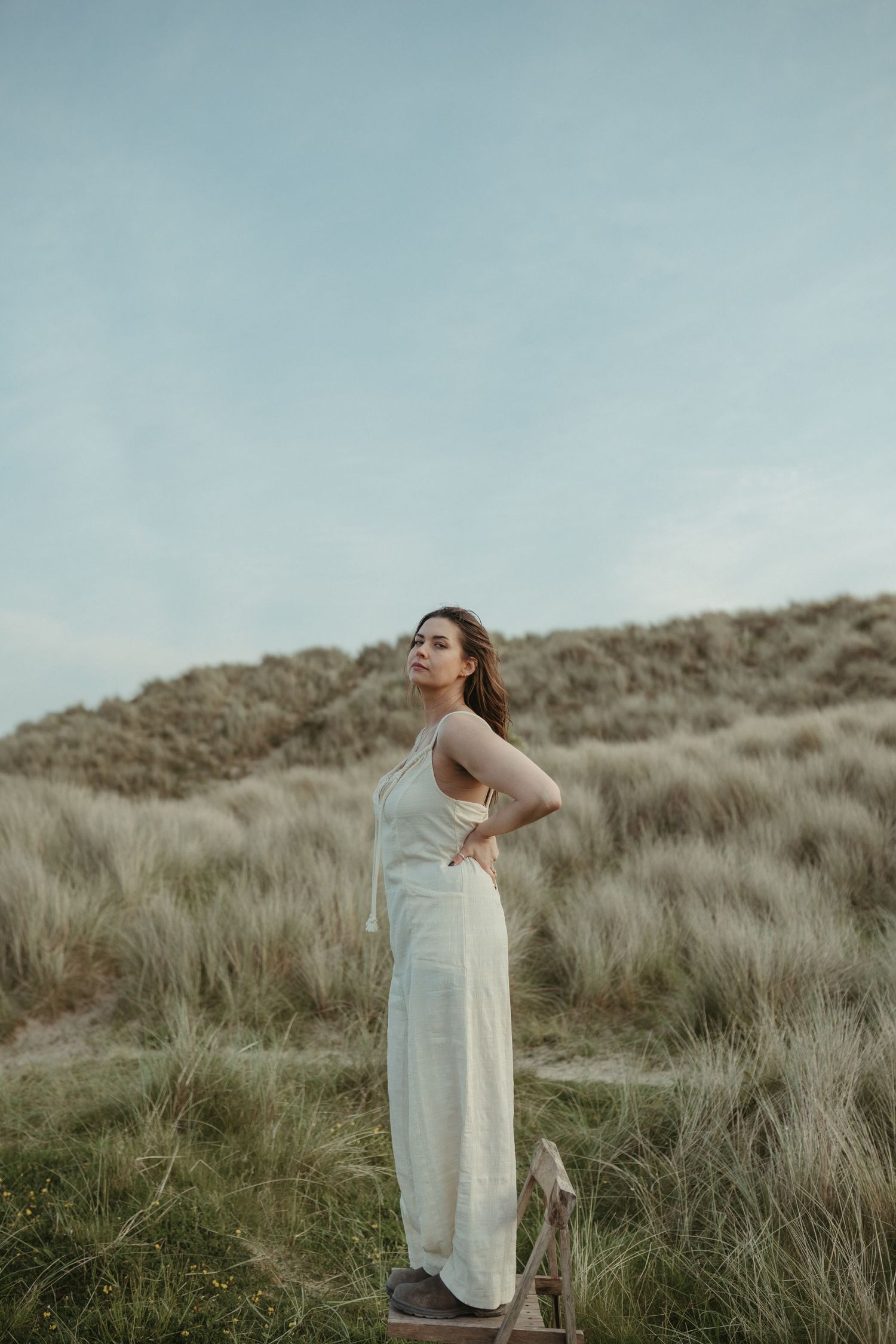 A white-dressed figure stands on a wooden platform amid windswept coastal grasses.