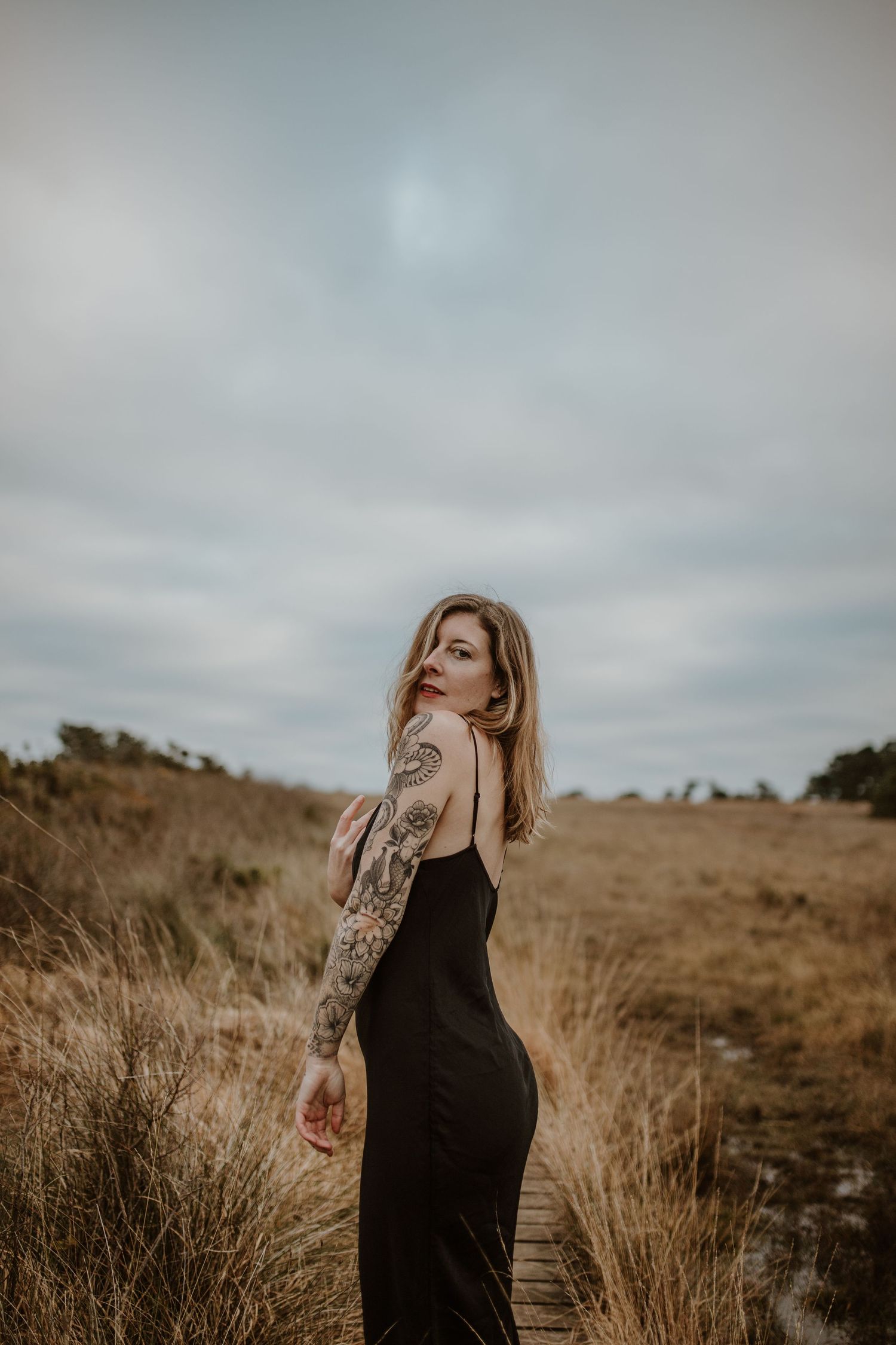 Someone poses in a black dress showing tattoo sleeve against a desert landscape backdrop.