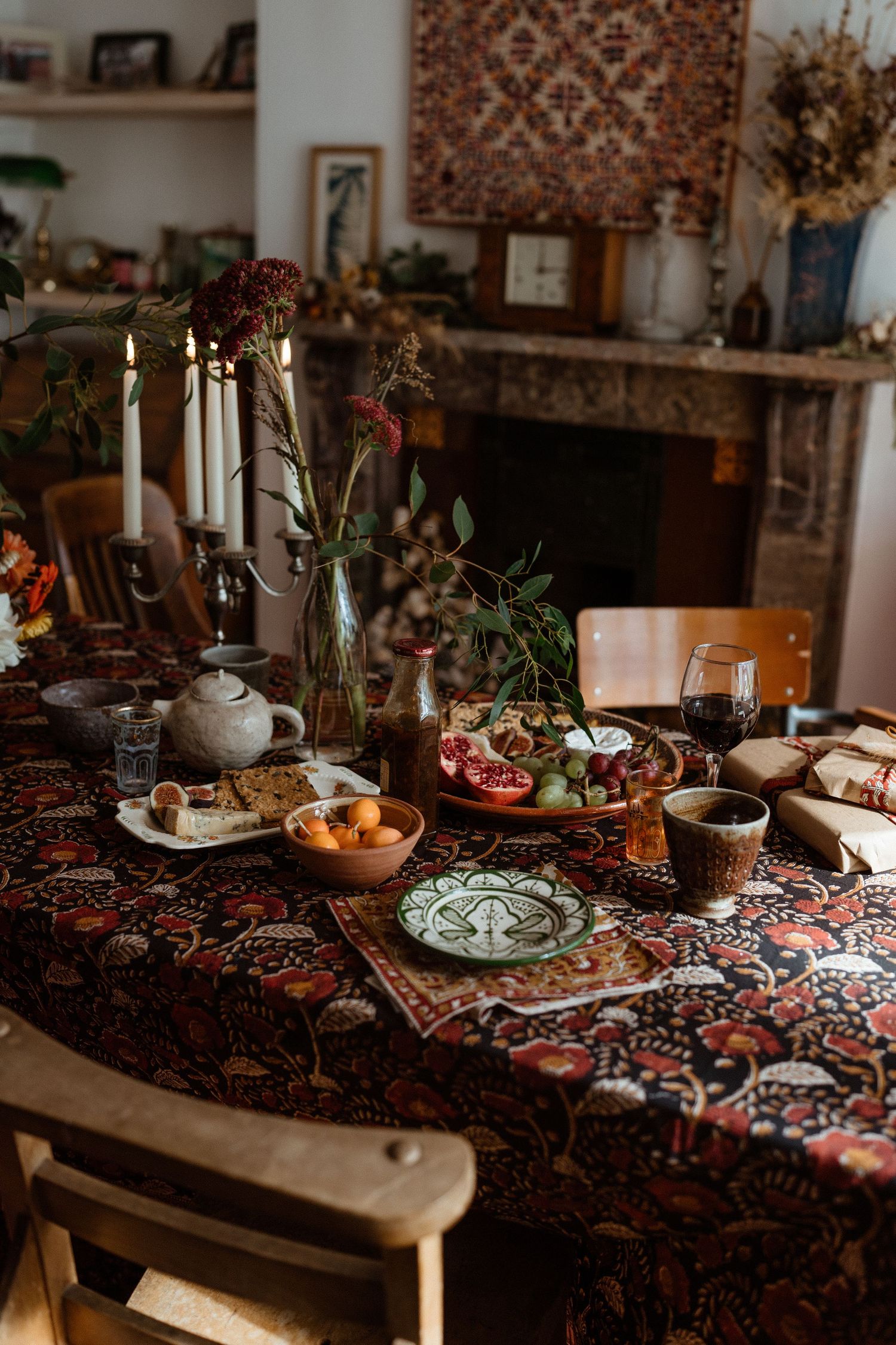 Cozy dining scene with candles, fruits and decorative plates on floral tablecloth near fireplace.