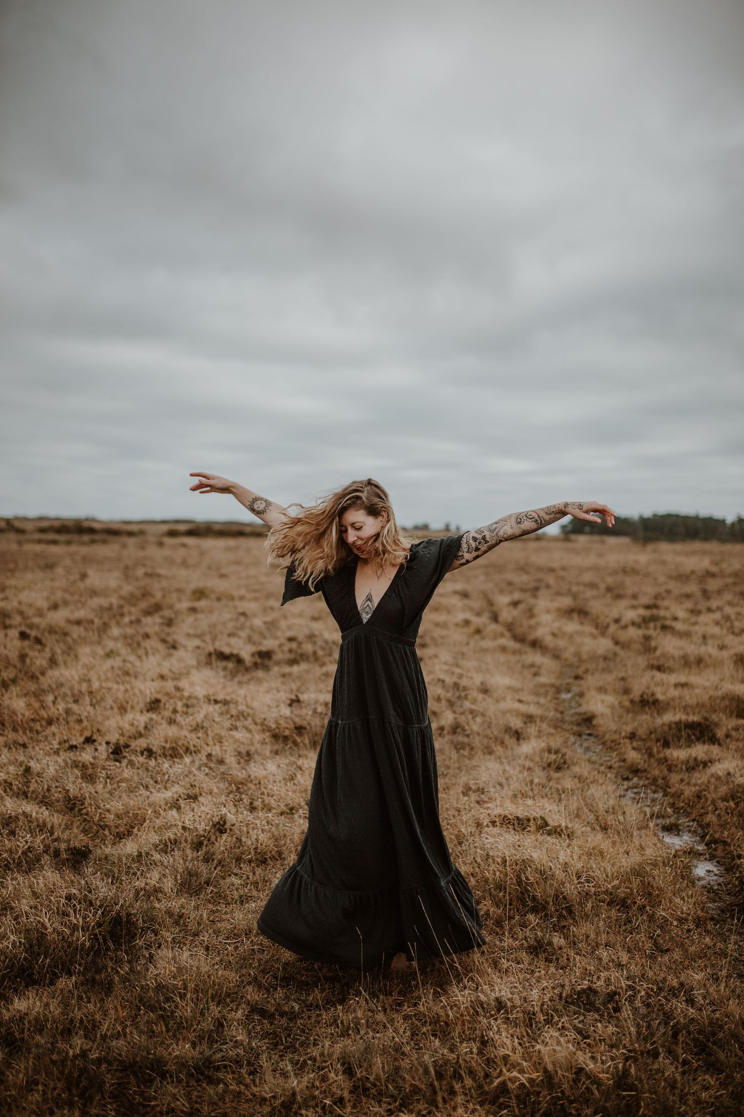 Someone in a flowing black dress dances with arms outstretched in an open field under grey skies.
