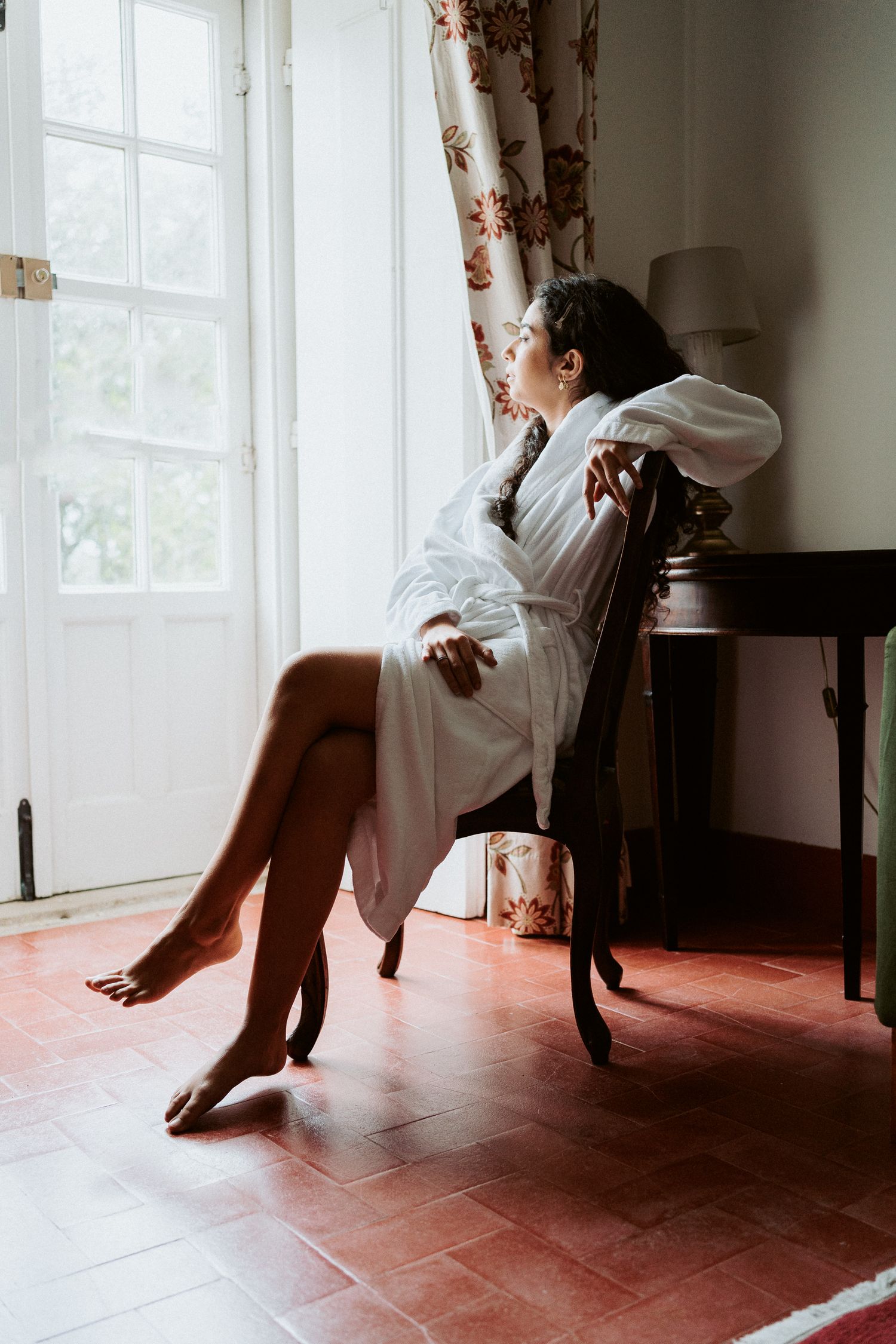 Artistic boudoir photo of someone in a white robe sitting by a window in morning light.