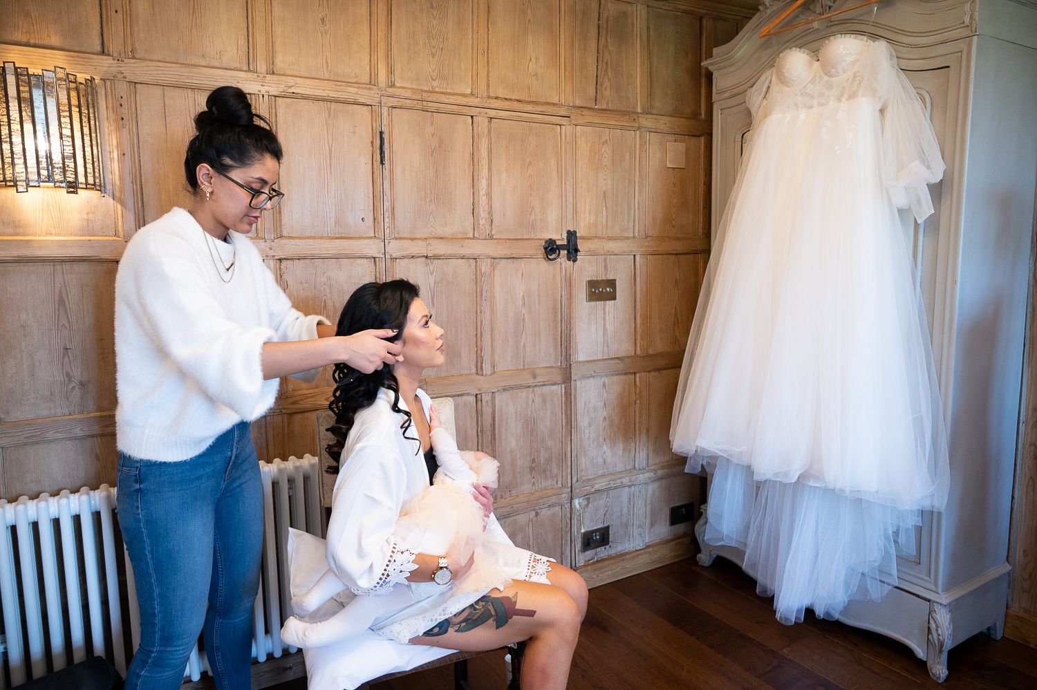 Wedding preparation scene showing a makeup artist styling a bride in a wood-paneled room with a dress hanging.