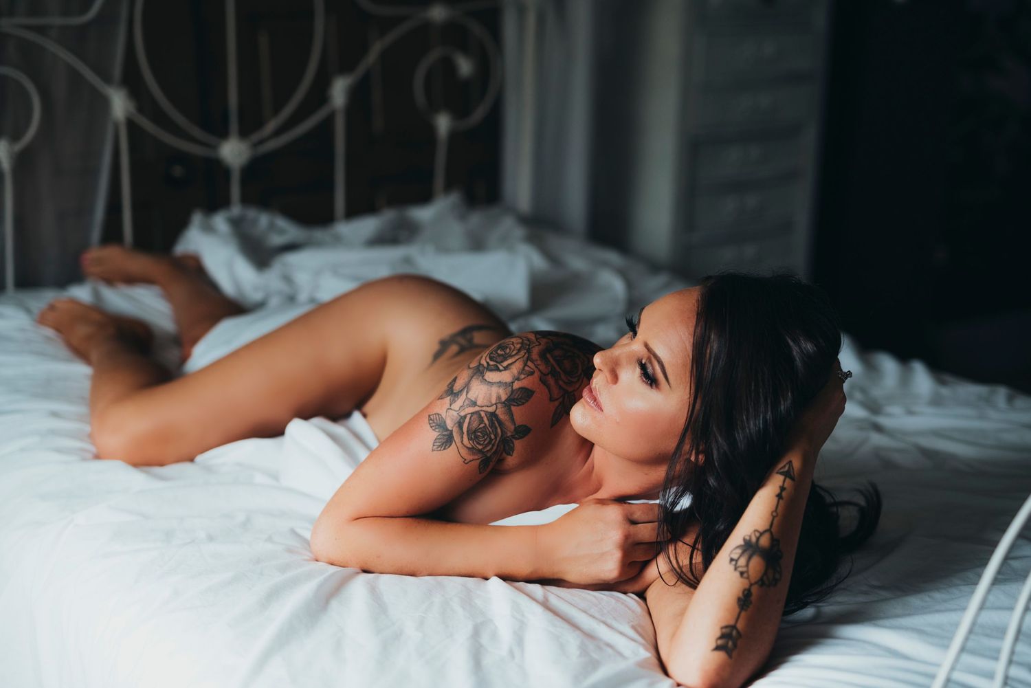 A tattooed person in minimal attire relaxes on white bedding in a moody boudoir setting.