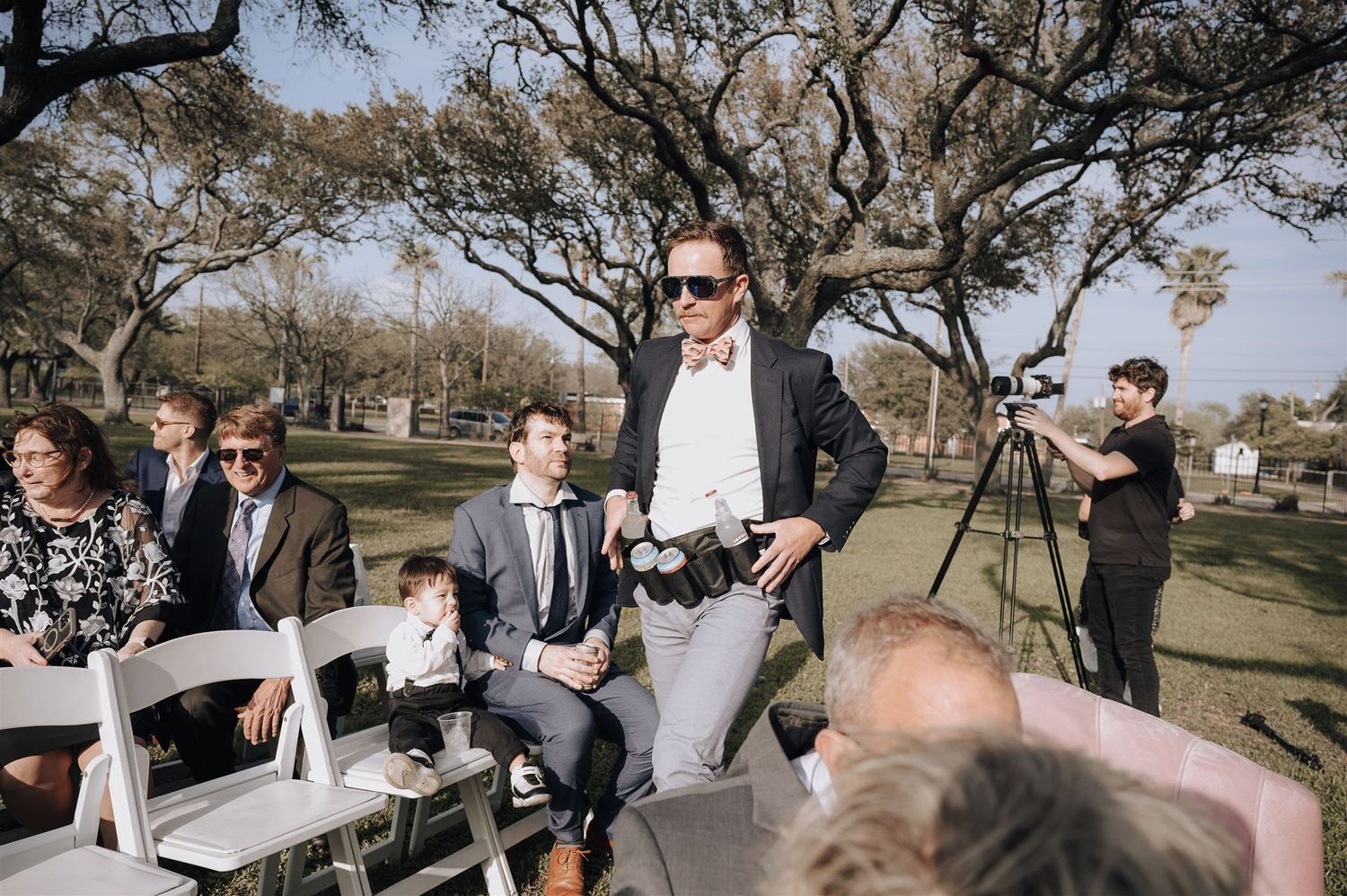 Wedding guests seated in white chairs under oak trees at an outdoor ceremony.