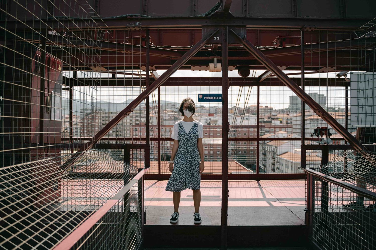 Person in polka dot dress standing on an industrial rooftop platform overlooking a cityscape.