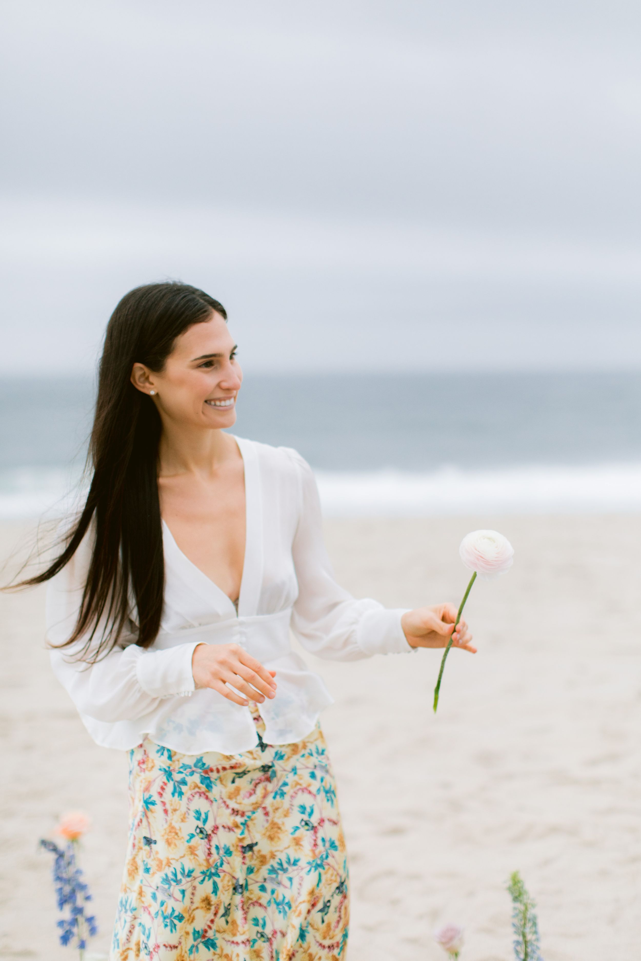 A woman in a floral dress poses playfully among flowers on a sandy beach.