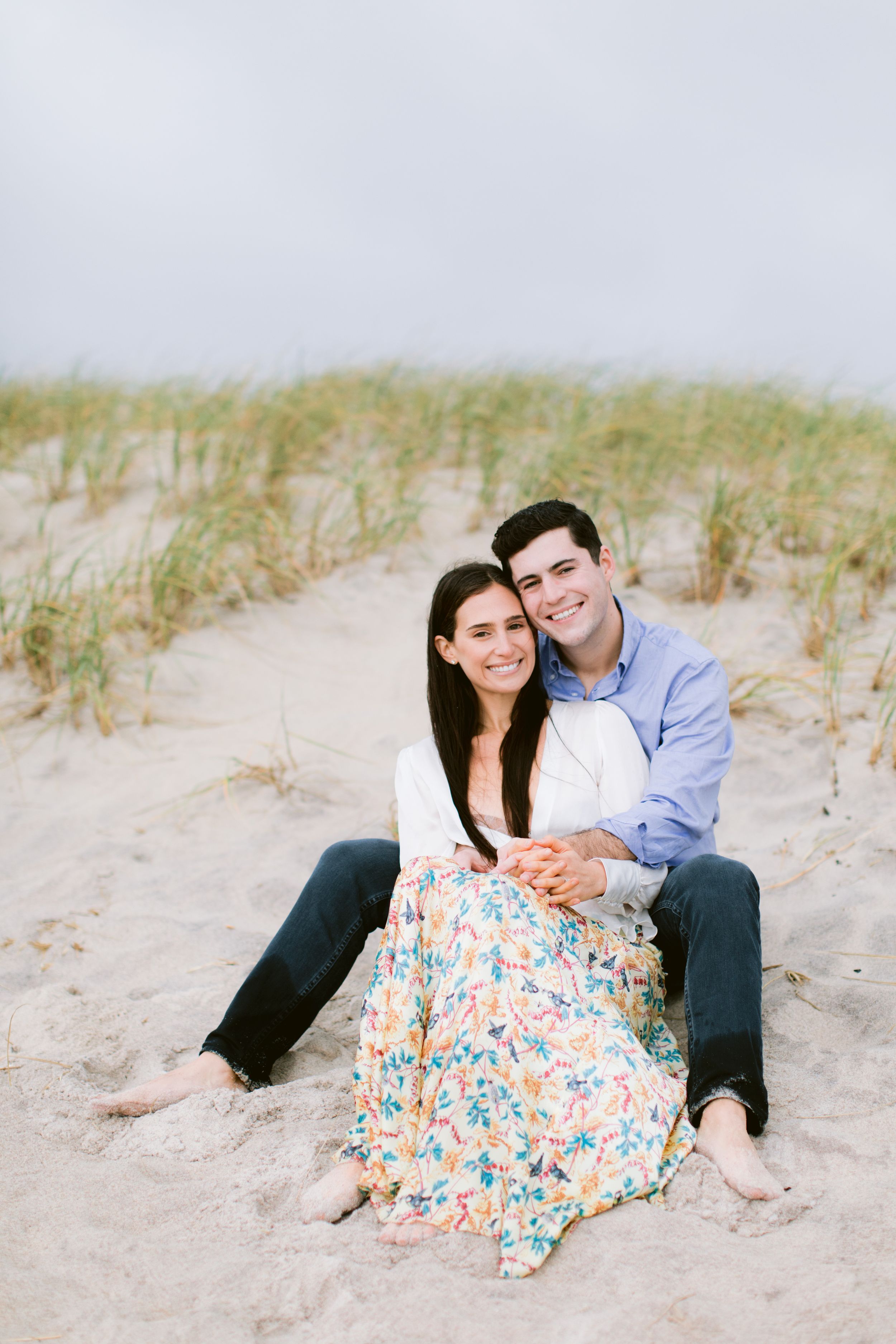 A couple relaxes together on sandy dunes wearing floral dress and blue shirt.