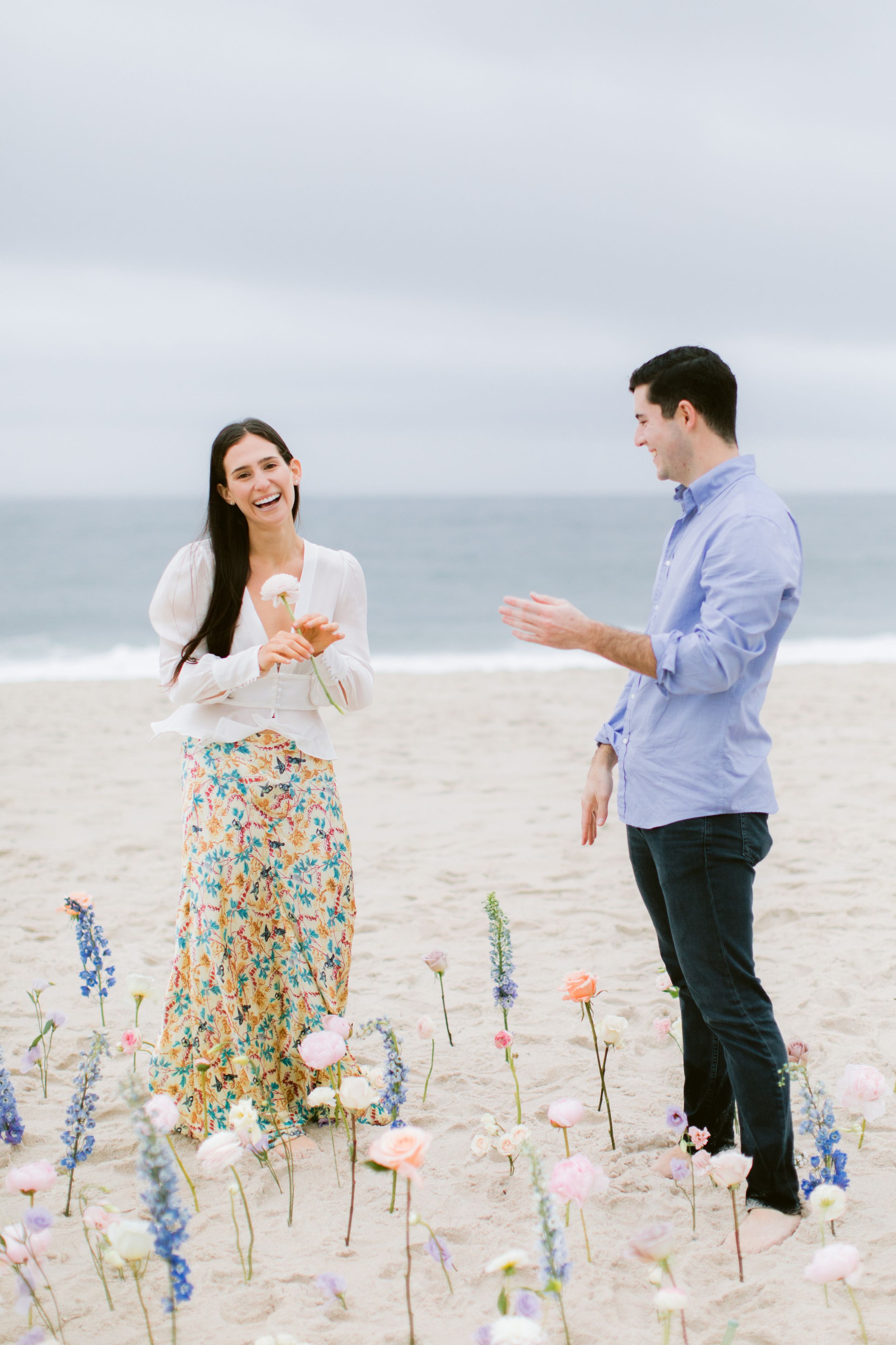 A couple interacts playfully among pink and blue flowers planted in beach sand.
