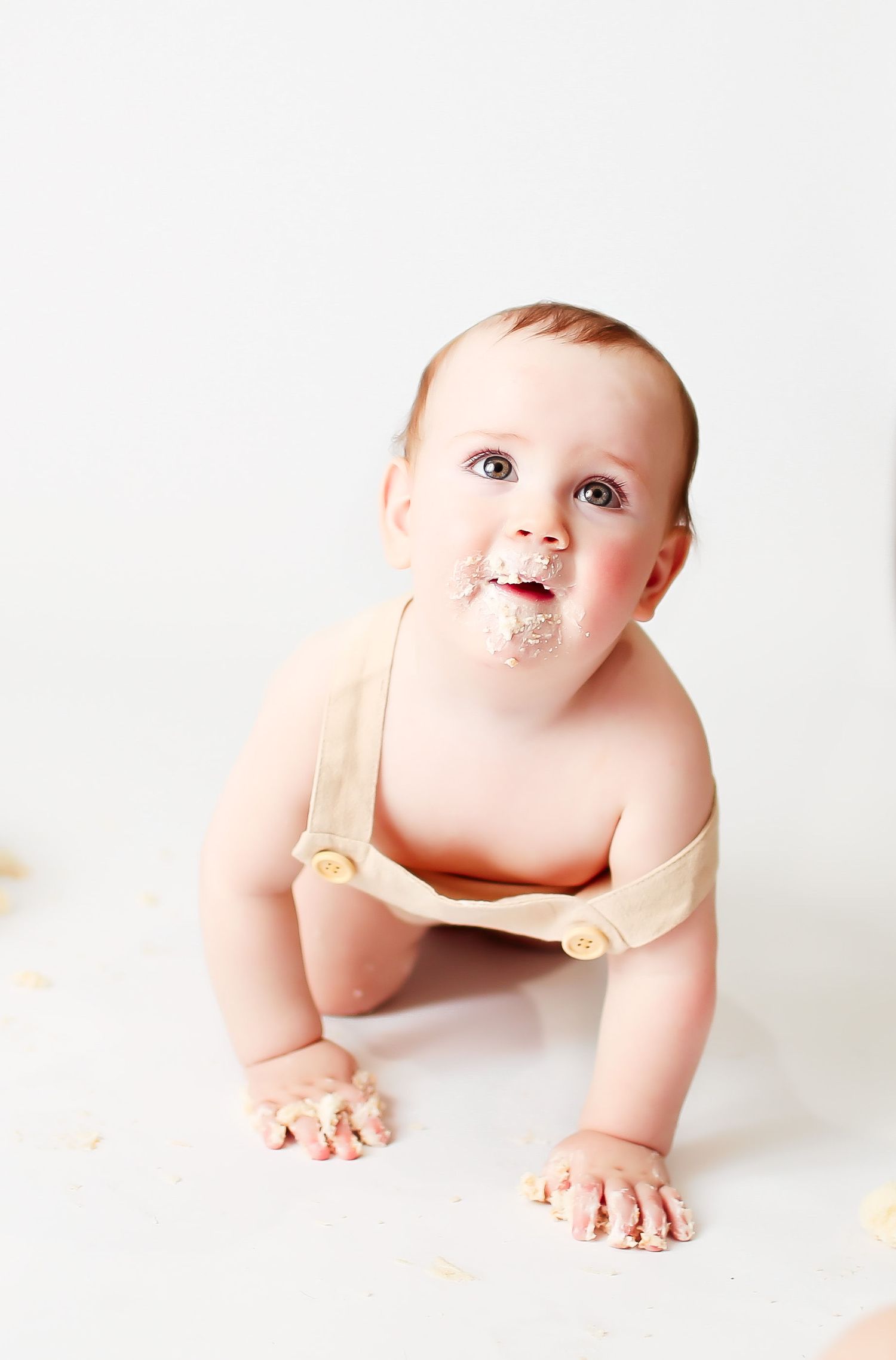 Adorable baby in beige suspenders crawling and smiling on white background with playful expression.