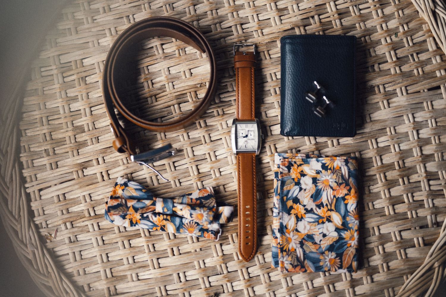 Wedding accessories including a leather belt, watch, wallet and floral tie laid on woven surface.