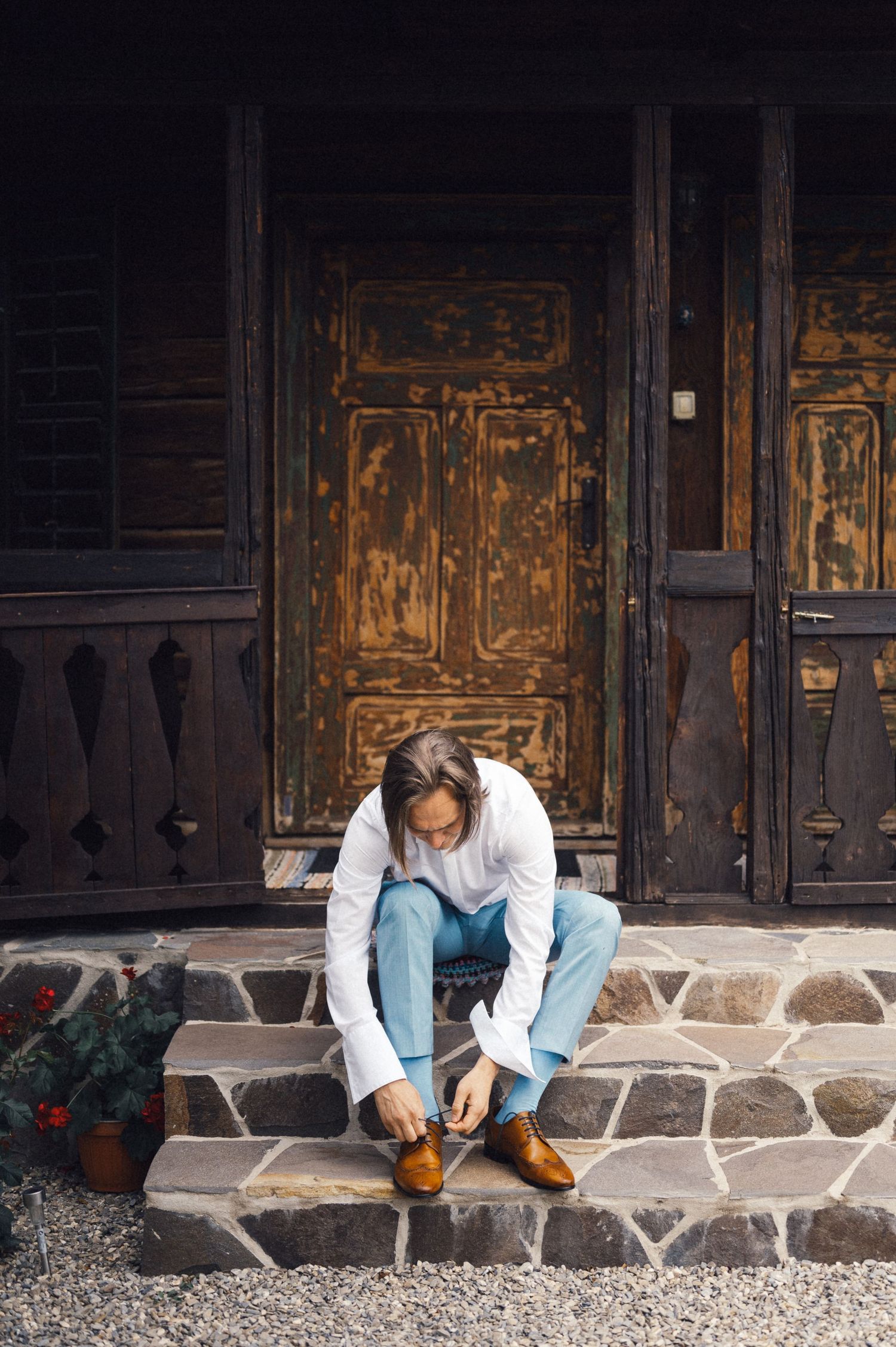 Someone sits on stone steps tying brown shoes in front of a rustic wooden door.