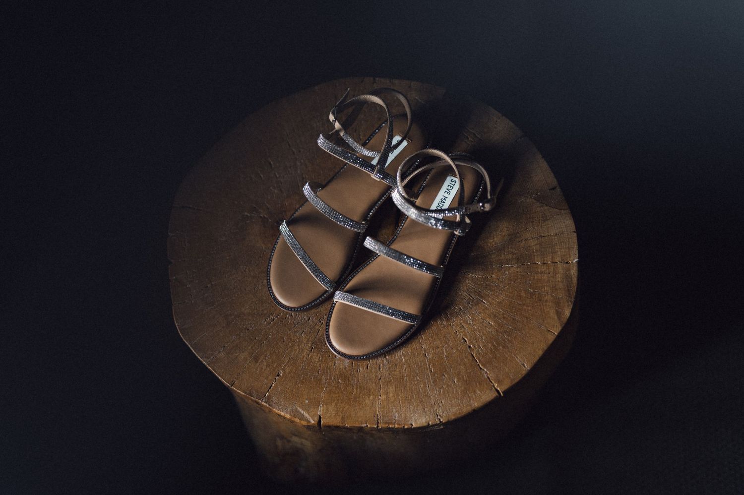 Silver sandals displayed on a round wooden surface against dark background.