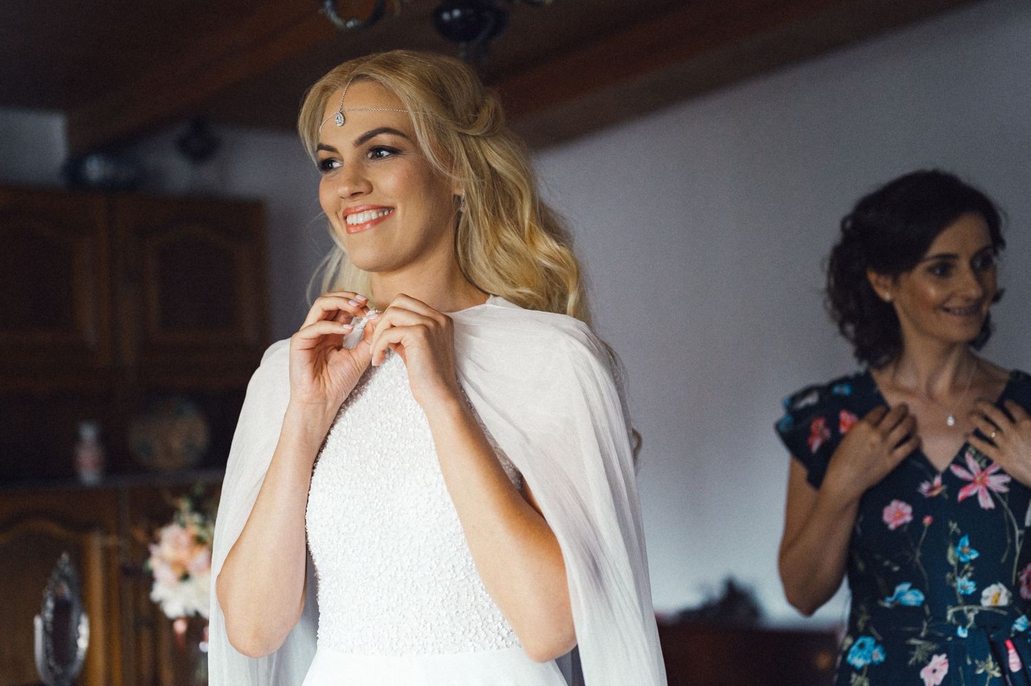 Bride in white dress and cape smiling while getting ready with help in floral dress.