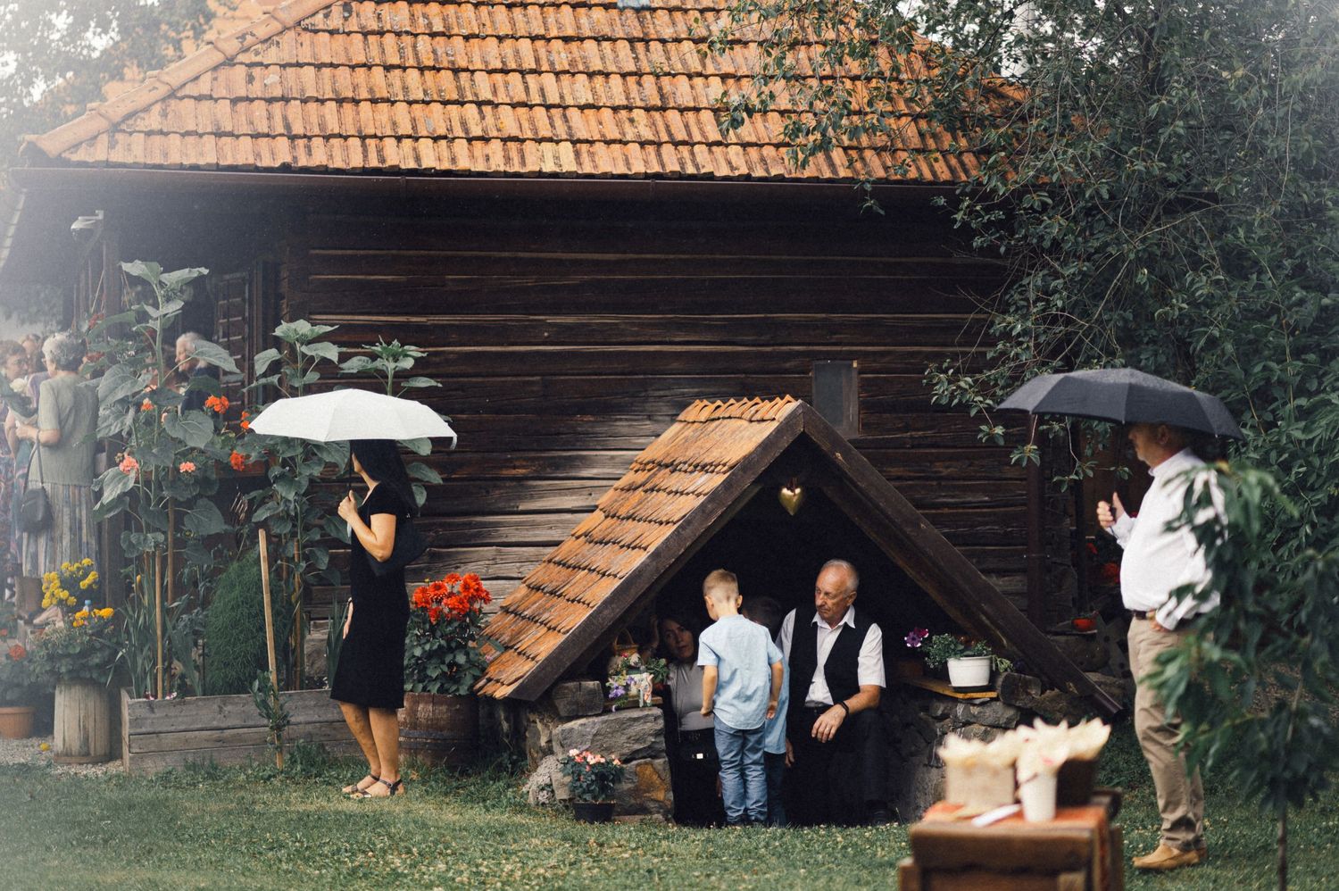 People with umbrellas gathering outside rustic wooden cabin in garden setting.