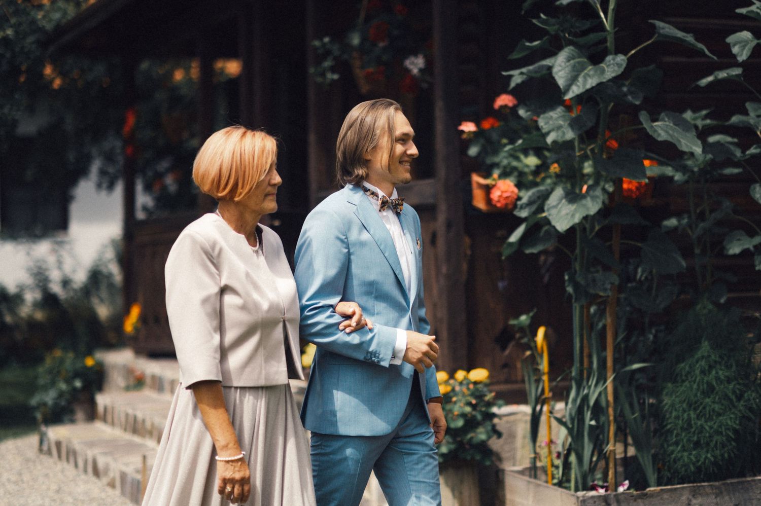 Two people in light colored outfits walking past flowering plants in a garden.