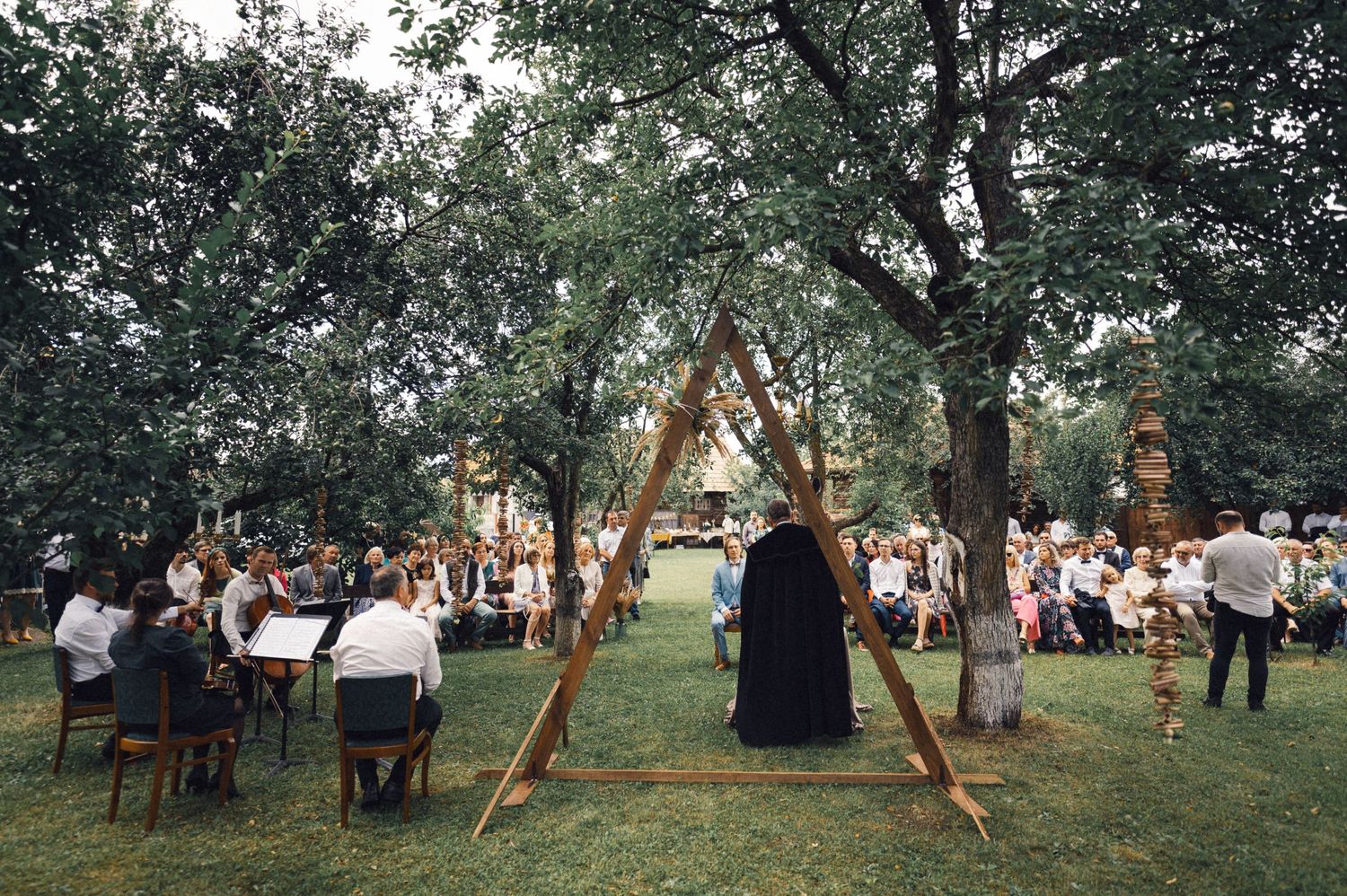 Outdoor ceremony setup with wooden chairs and triangular arch structure under shady trees.