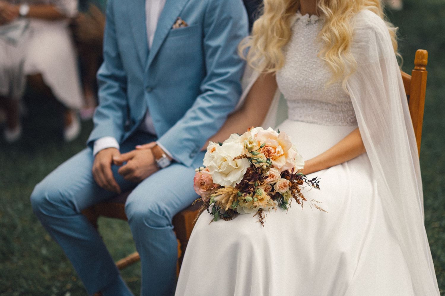 A couple in wedding attire sits outdoors with a peach and white floral bouquet.