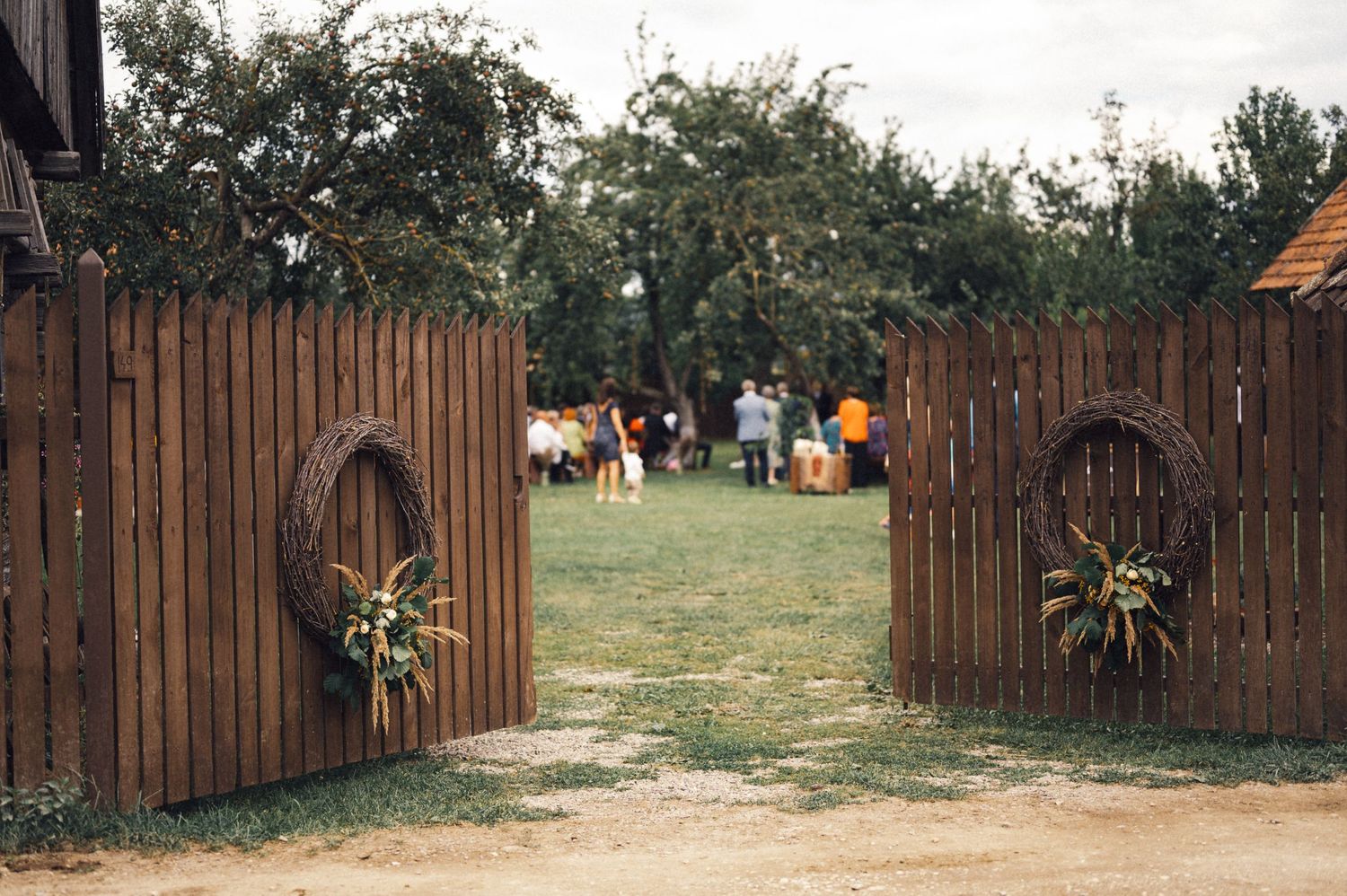 A rustic wooden gate entrance decorated with floral arrangements leads to an outdoor gathering.