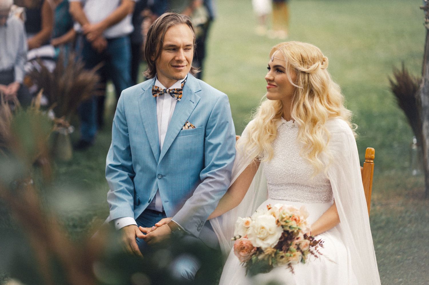 A couple in a light blue suit and white wedding dress stands during an outdoor ceremony.