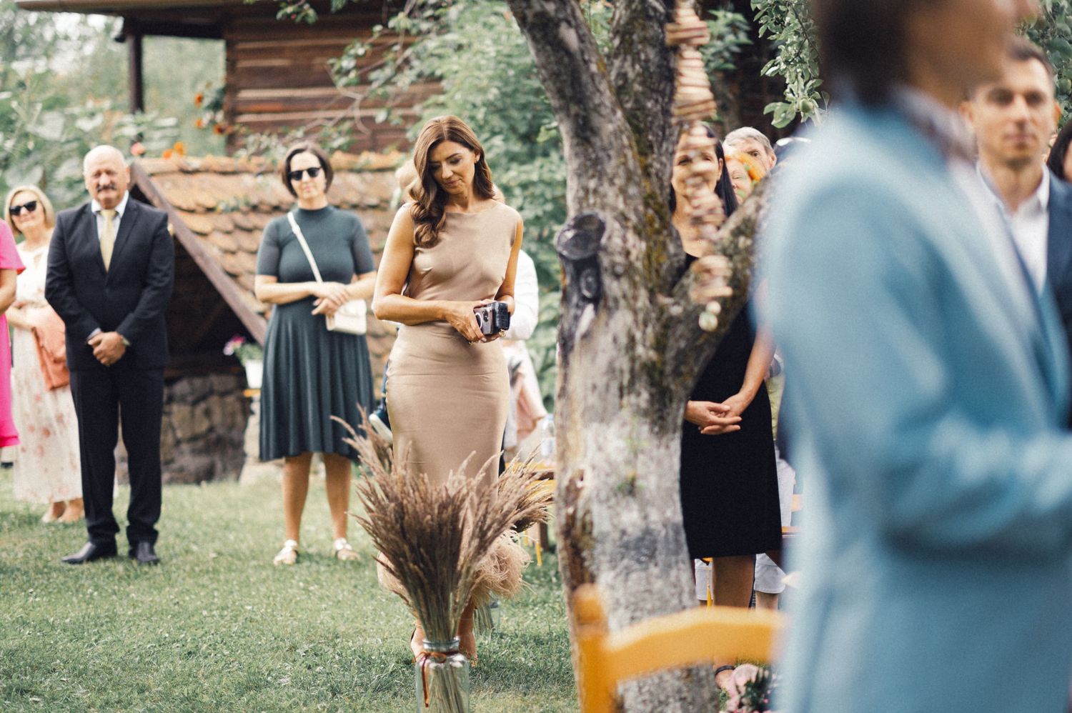 Wedding guests gather on a lawn with wooden chairs and trees during an outdoor ceremony.