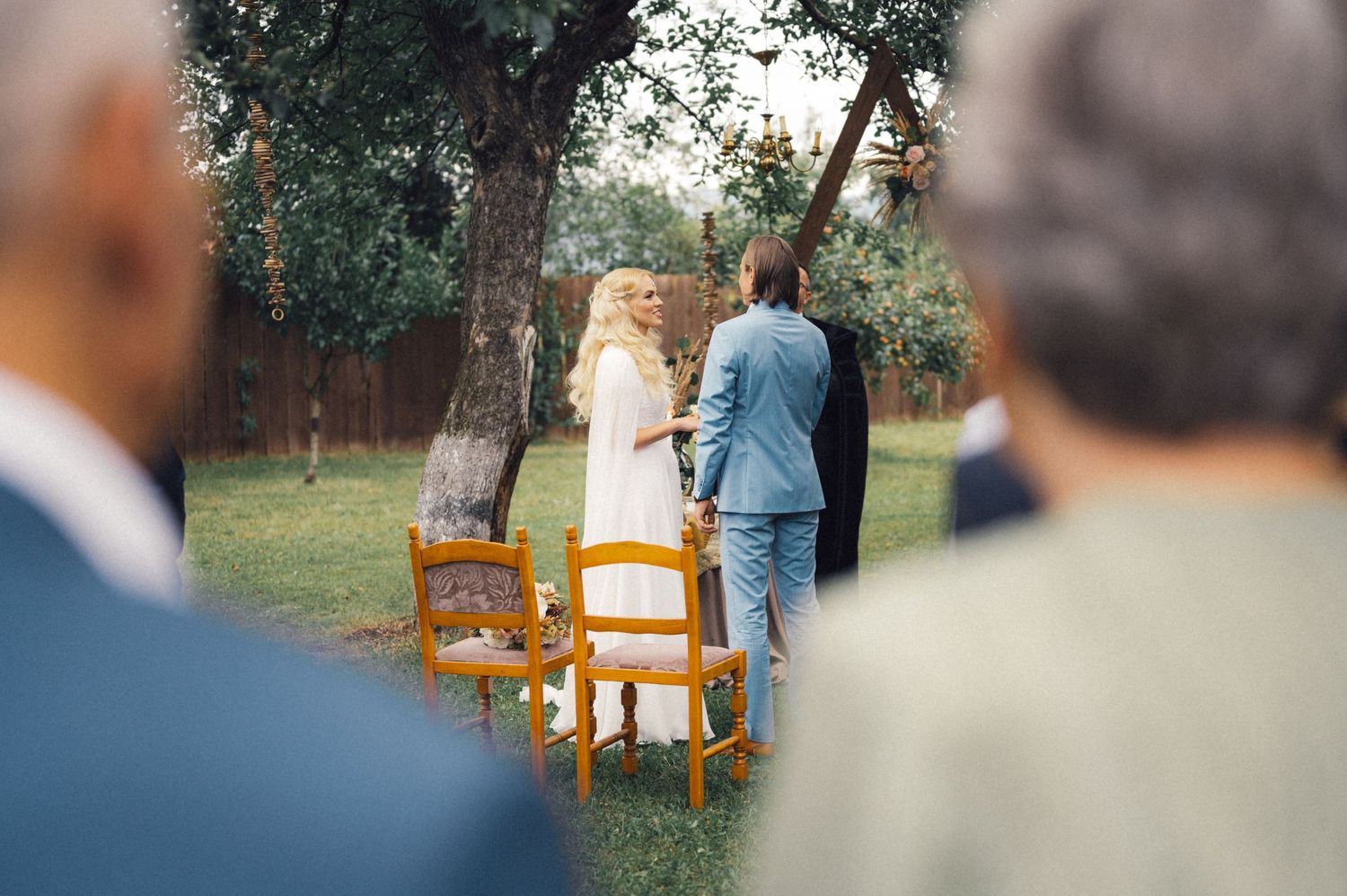 Guests watch as a couple stands near wooden chairs during an outdoor wedding ceremony.
