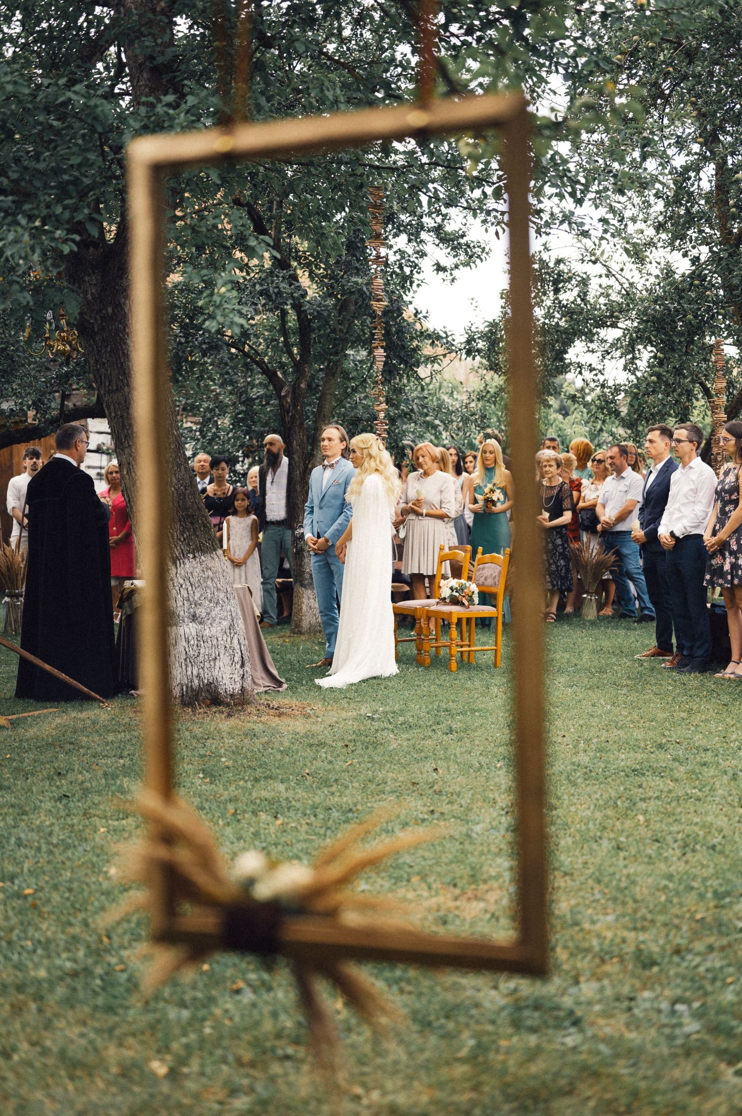 An artistic framed shot of a wedding ceremony through a golden frame in a garden setting.