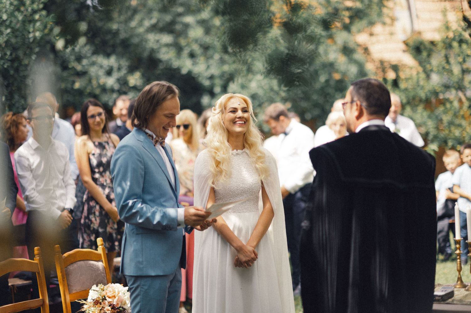 A couple stands at a rustic outdoor ceremony with an officiant in a garden setting.