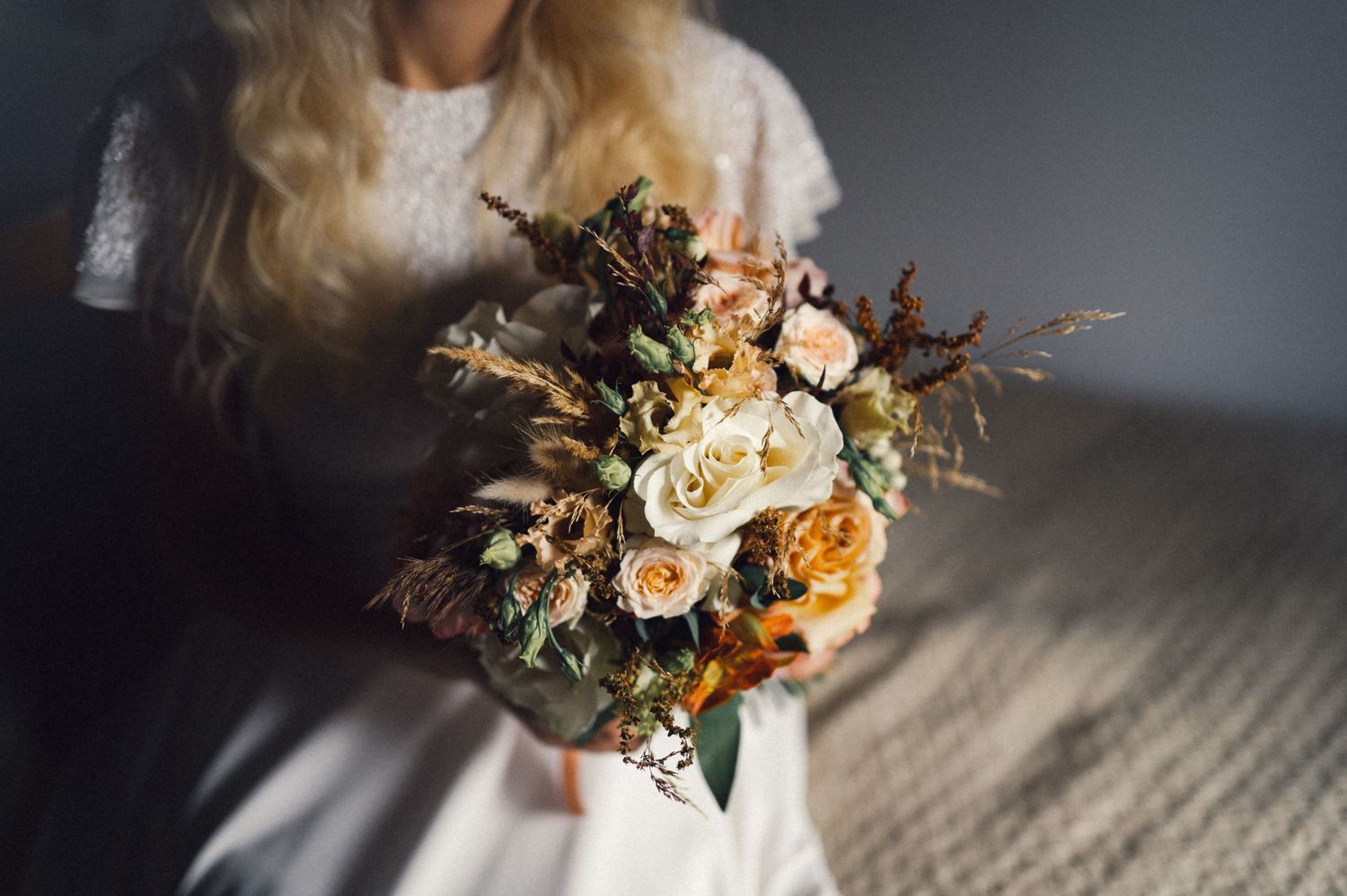 A beautiful autumn-toned wedding bouquet with roses and dried flowers held against a white dress.