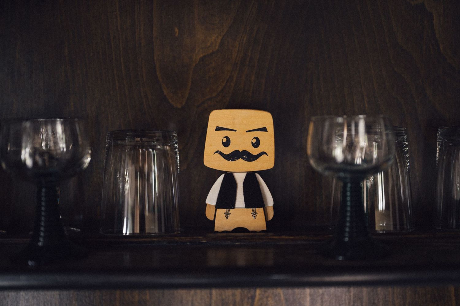 A wooden decorative figure with a mustache displayed between wine glasses on a dark shelf.