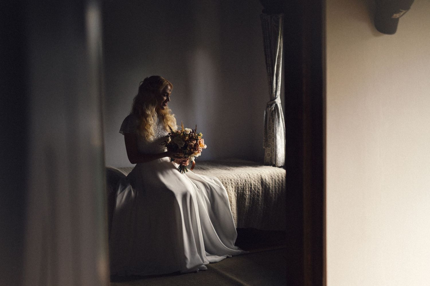 Silhouette of a bride holding flowers while sitting in dramatic window light.