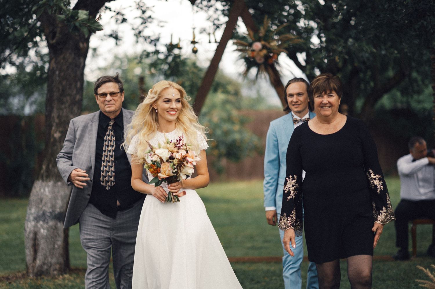 A bride in white walks through a garden setting accompanied by wedding guests.