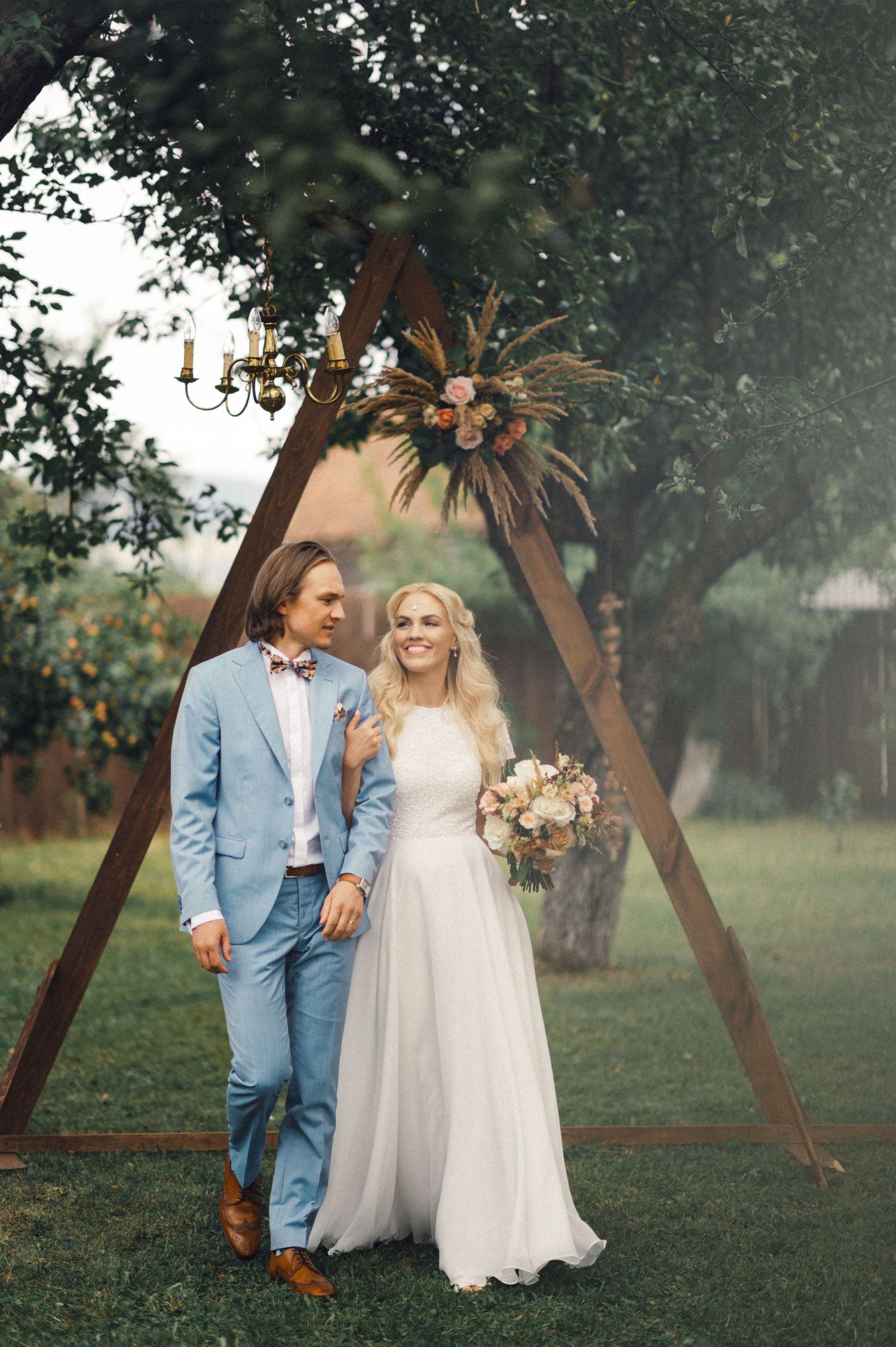 Newlyweds stand beneath a wooden triangle arch decorated with flowers in a garden setting.
