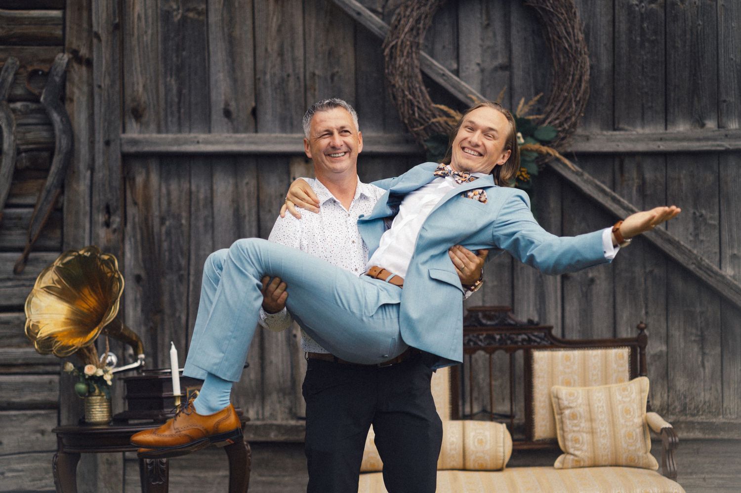 A playful wedding photo with groom carrying bride in front of rustic barn backdrop.