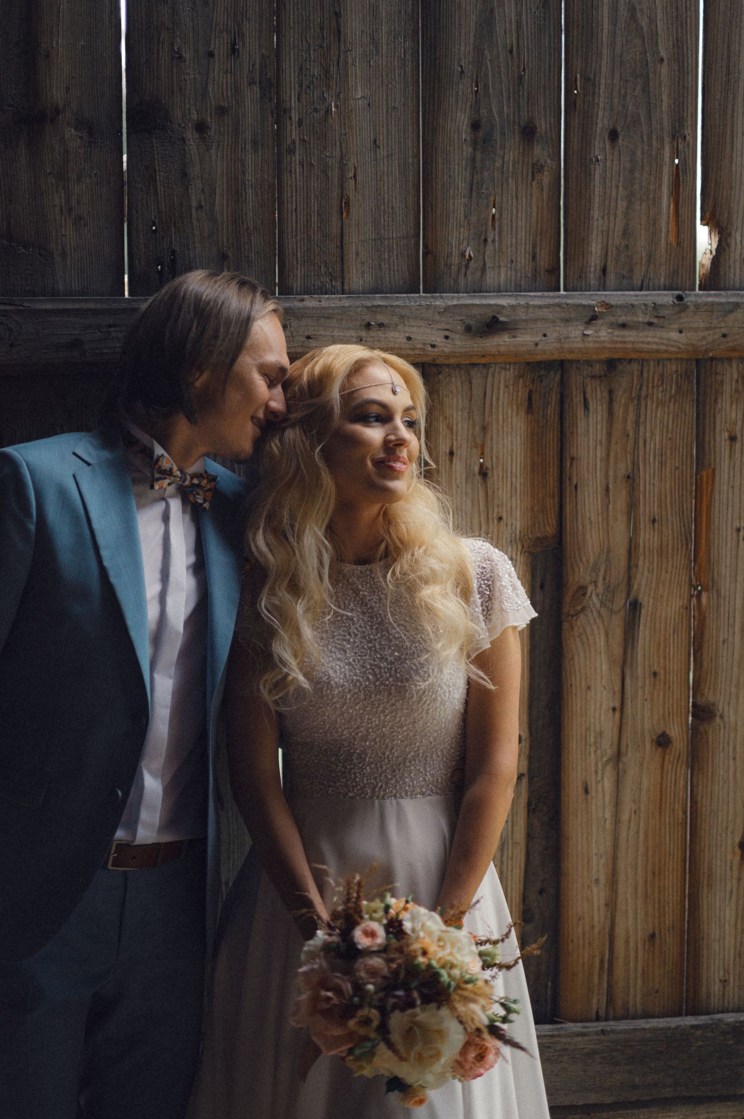 A romantic wedding couple poses against a rustic wooden barn wall with the bride holding a bouquet.