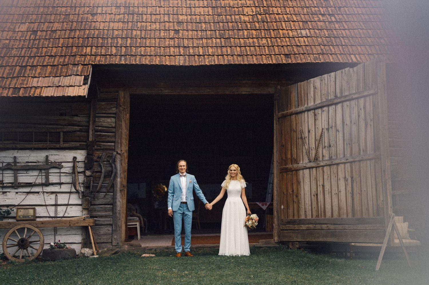 A couple stands in the doorway of a rustic barn holding hands during their wedding day.