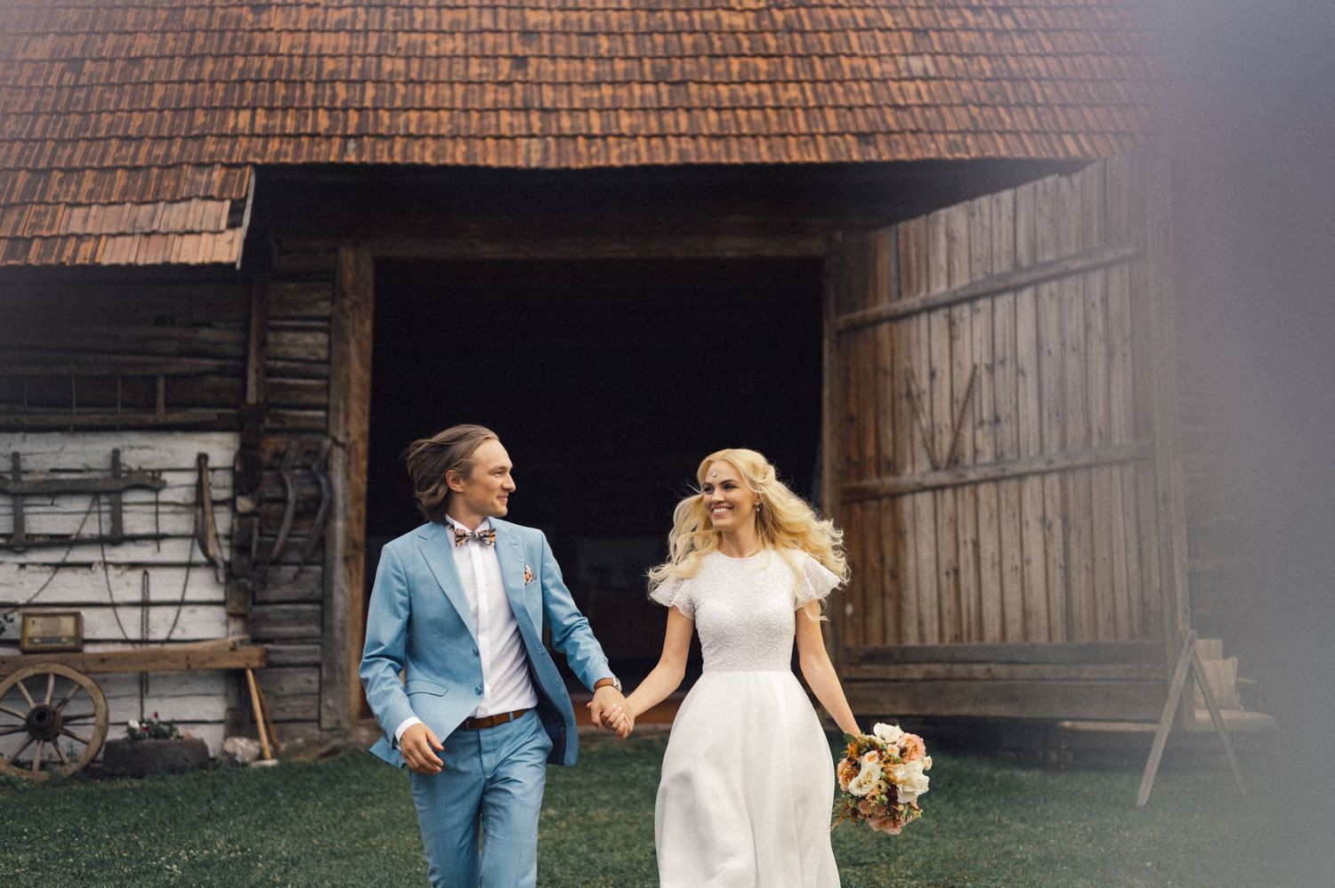 A couple walks hand in hand outside a rustic barn in casual wedding attire.