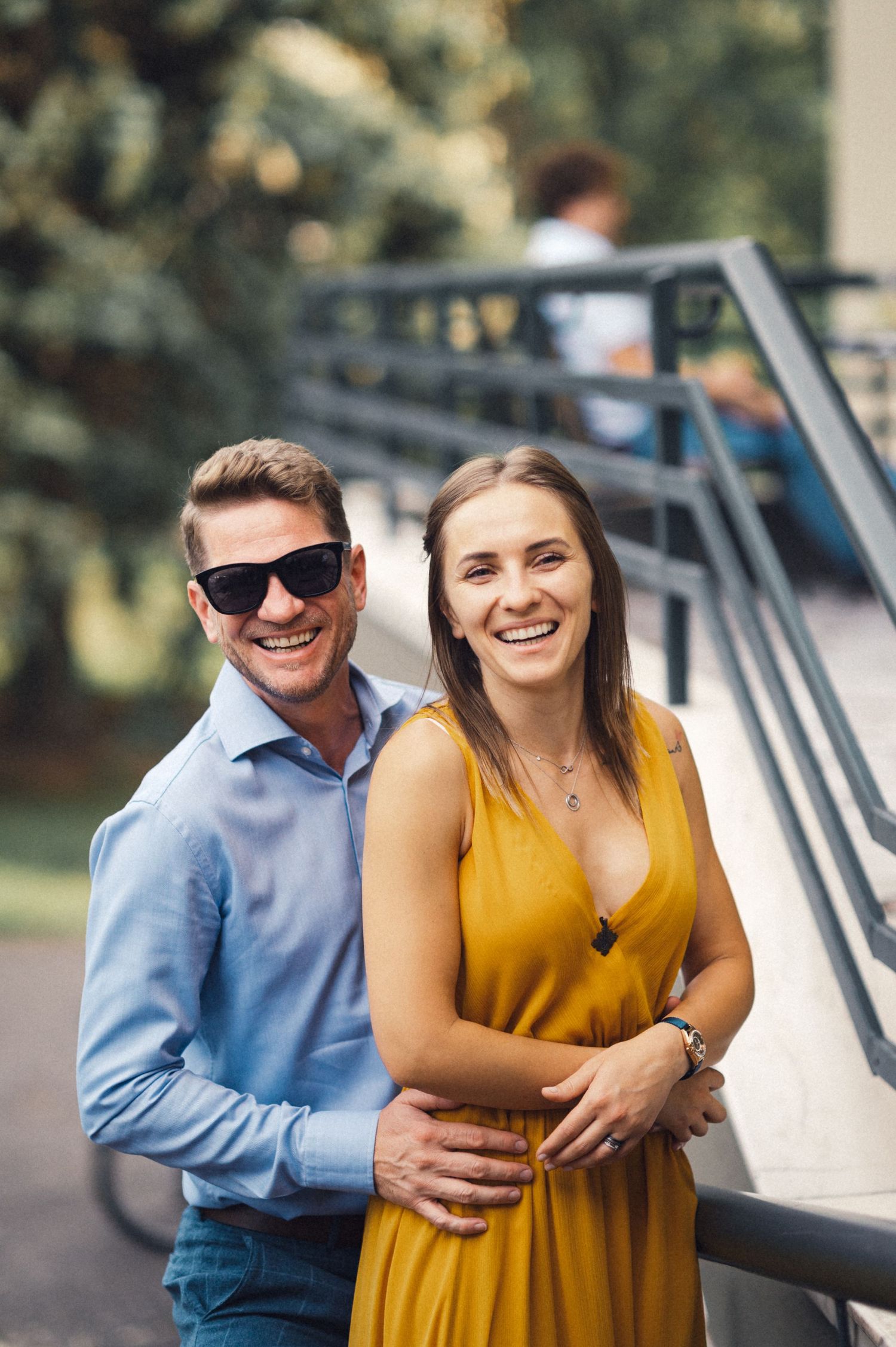 Happy couple embracing by metal railing outdoors, with her in yellow dress and him in blue shirt.