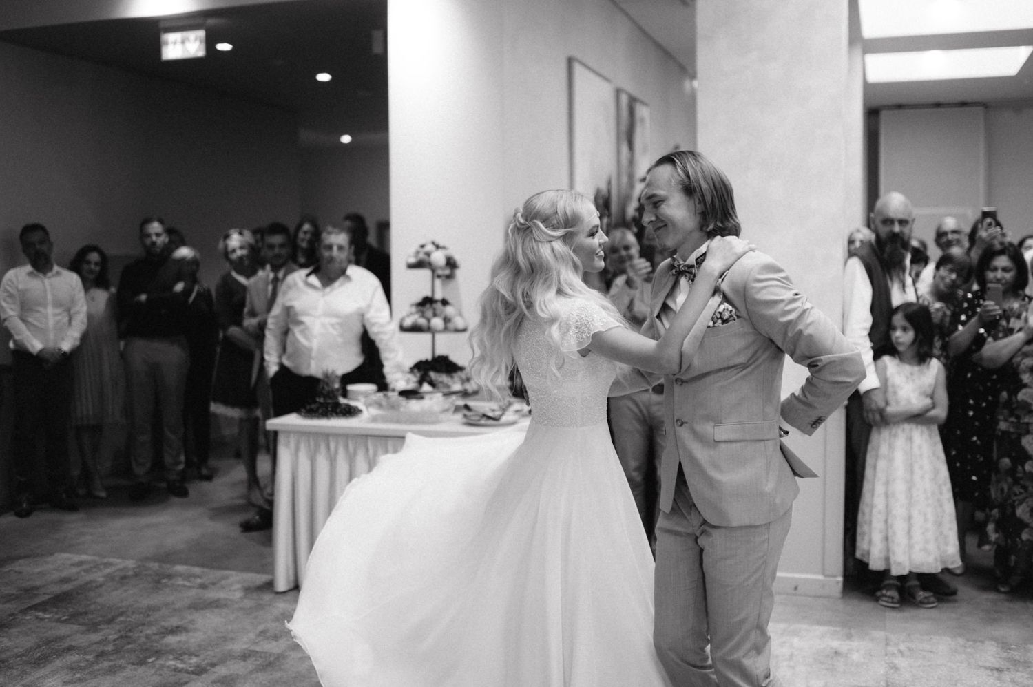 Black and white photo of couple dancing at wedding reception with guests in background.