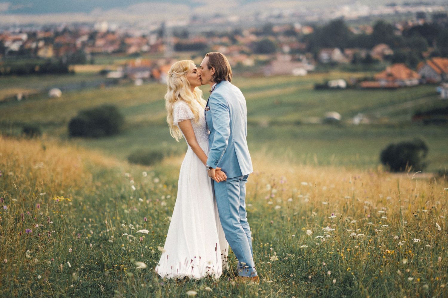 Couple embracing in a sunlit meadow with countryside views in the background.