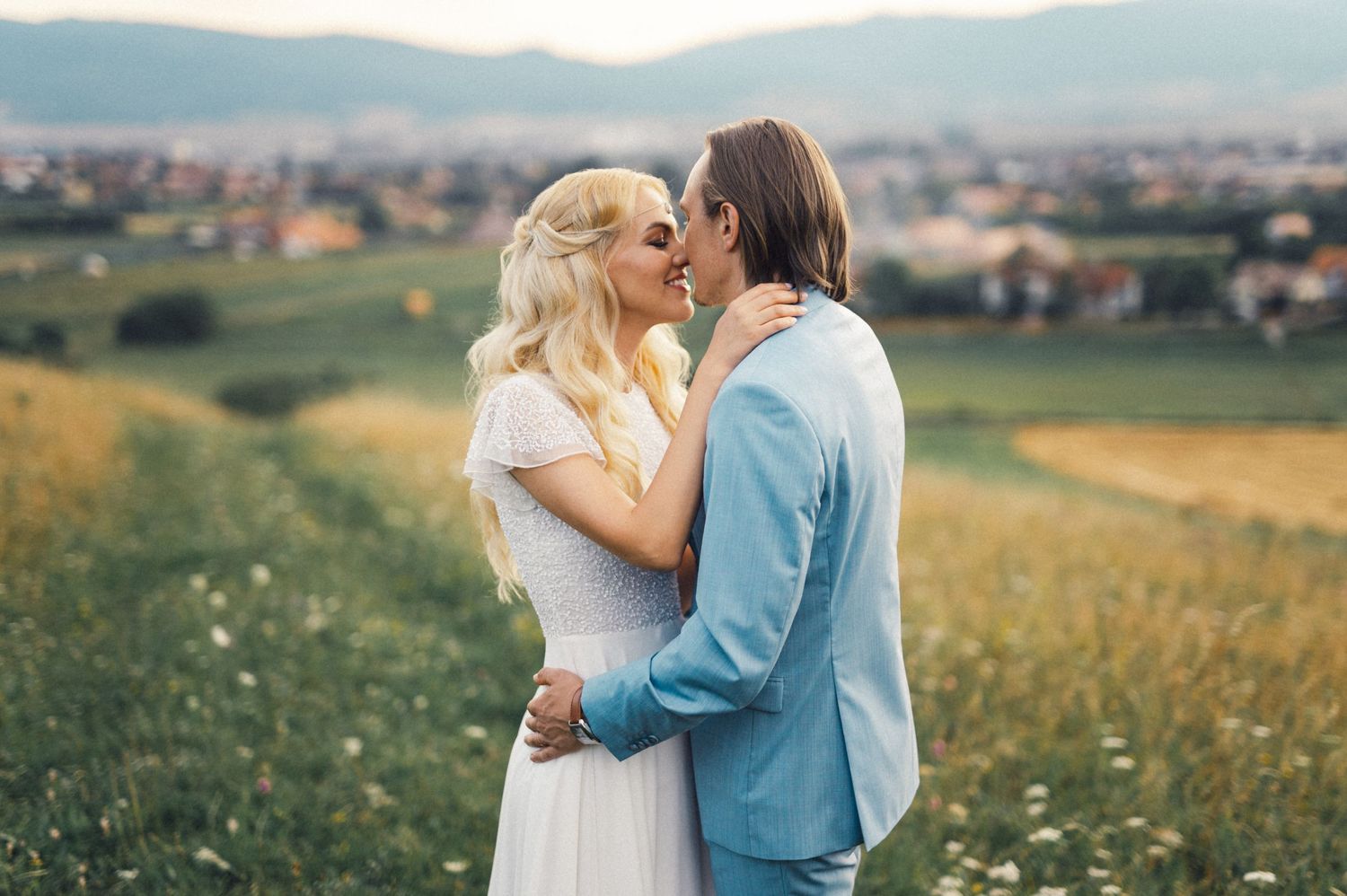 Romantic couple embracing in a meadow with rolling hills and city in background.