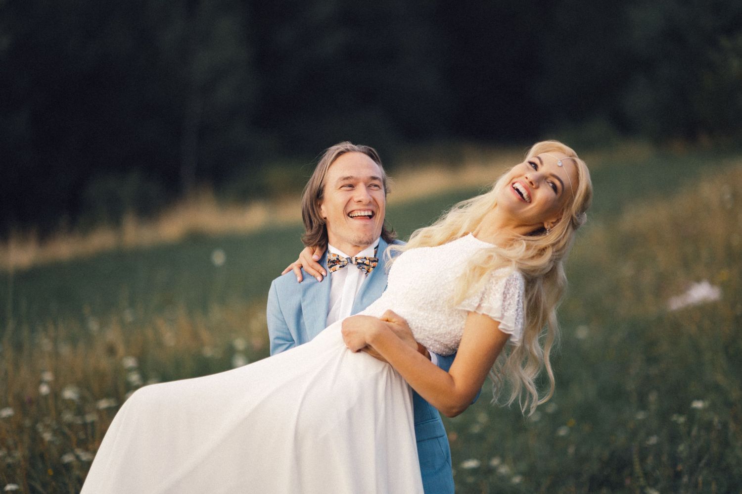 A joyful couple in a meadow share a romantic moment as he lifts her in a white dress against a scenic landscape.