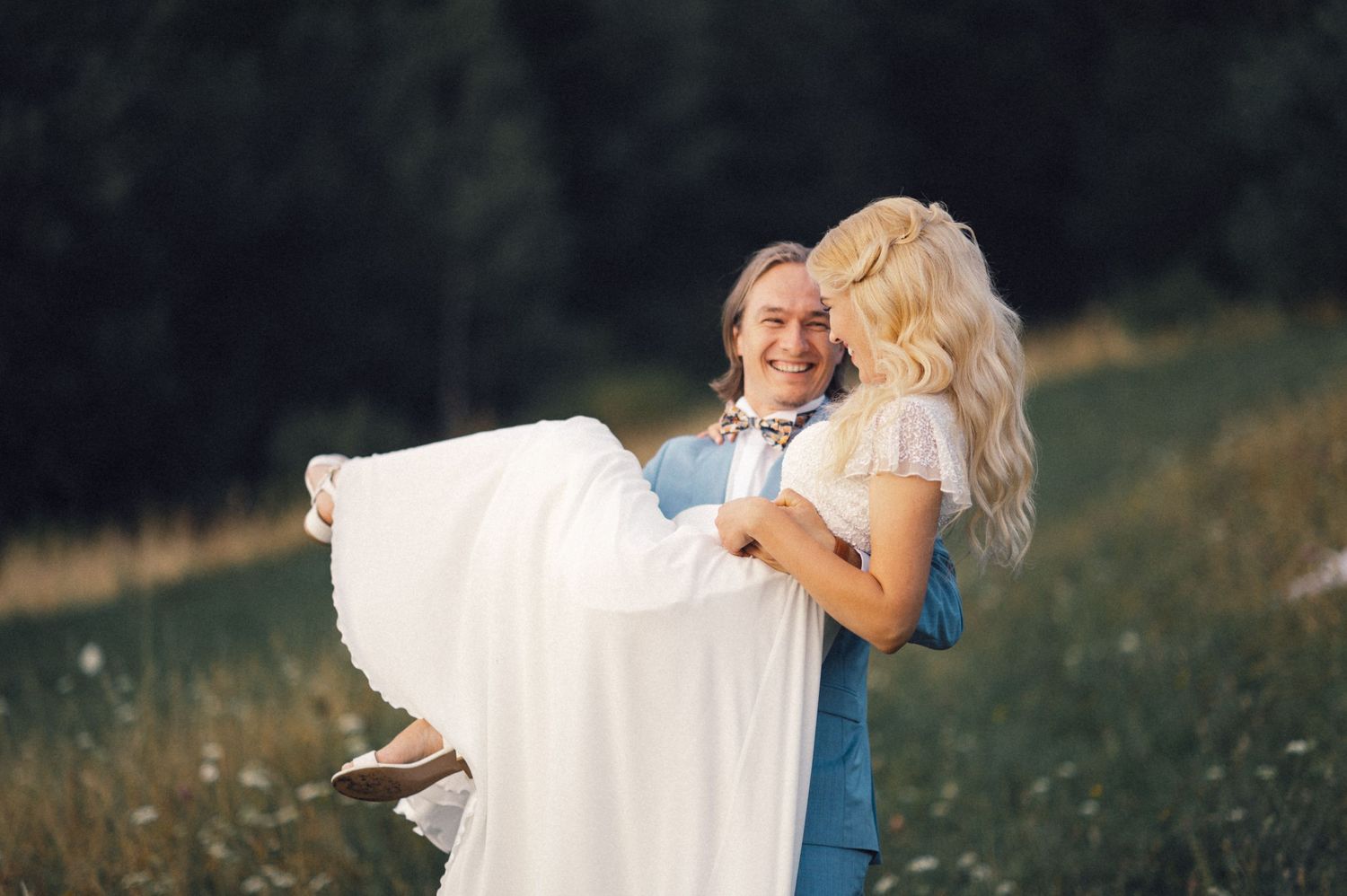 A couple shares a playful moment as he carries her in her flowing white dress in a sunny meadow.