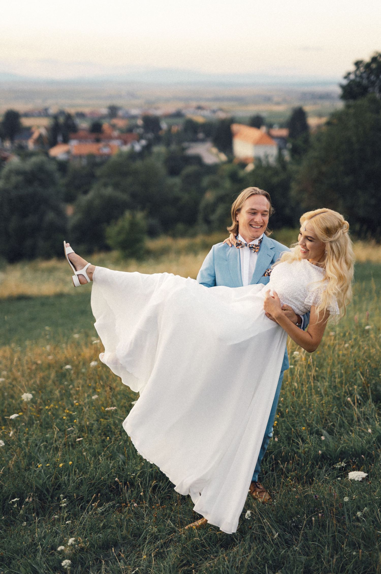 A couple poses on a hilltop overlooking a village as he lifts her in her flowing white dress.