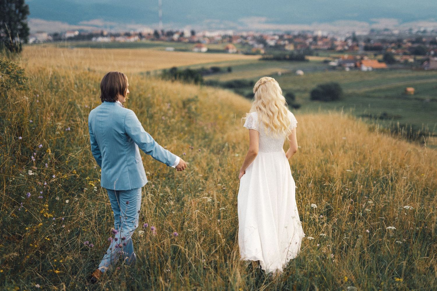 A couple in coordinated blue and white outfits walk through a scenic hilltop meadow with a village in the distance.