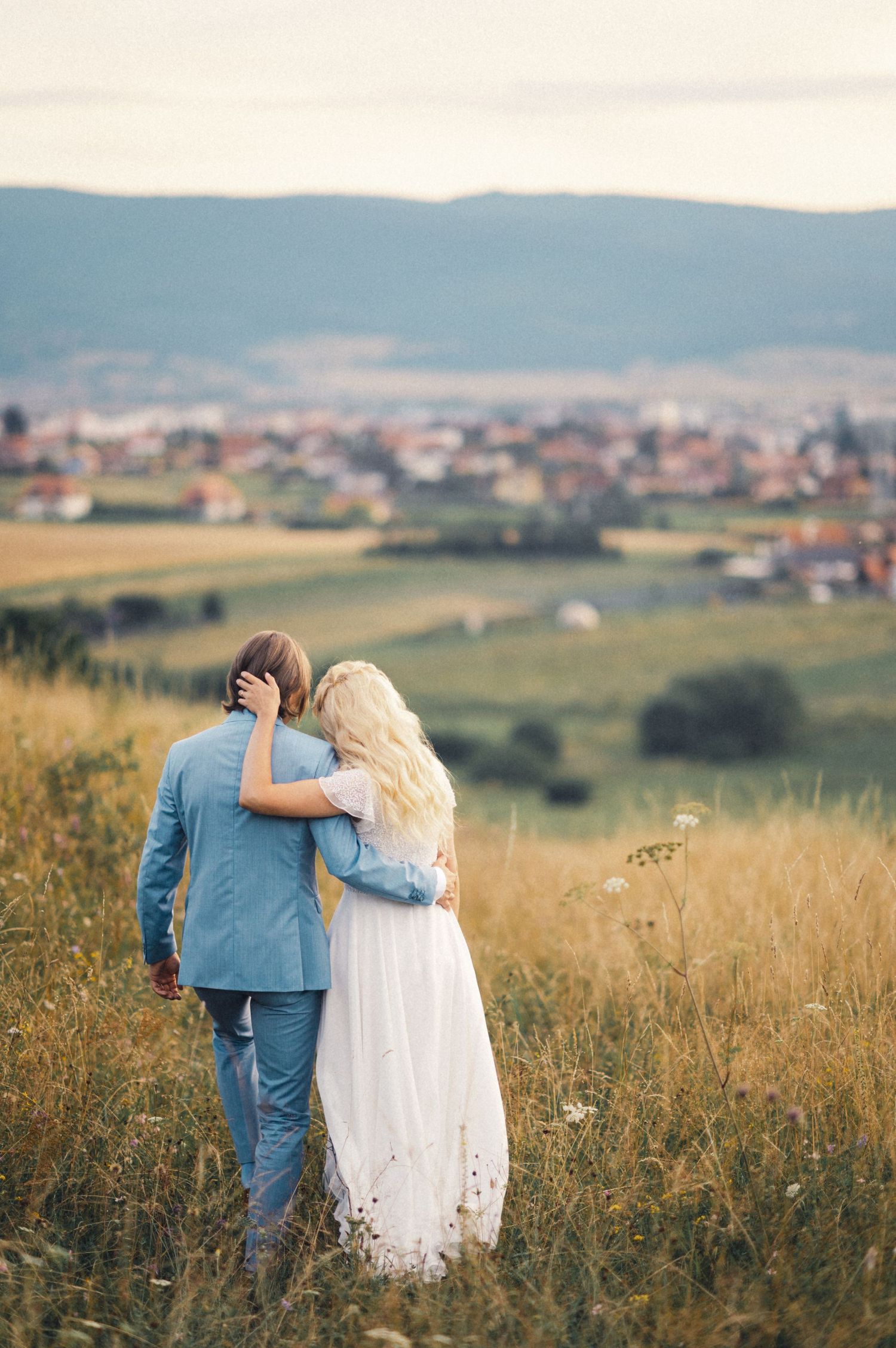 A couple embraces while looking out over a scenic rural landscape with rolling hills and distant houses.