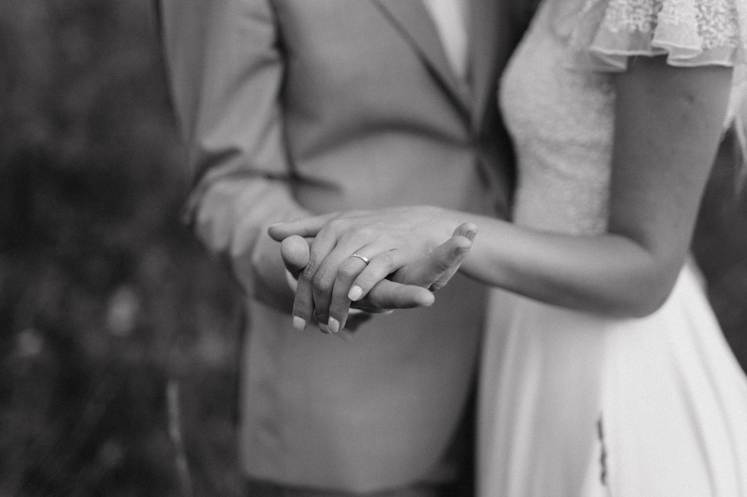 Black and white detail shot of newlyweds holding hands, showing wedding rings and formal attire.