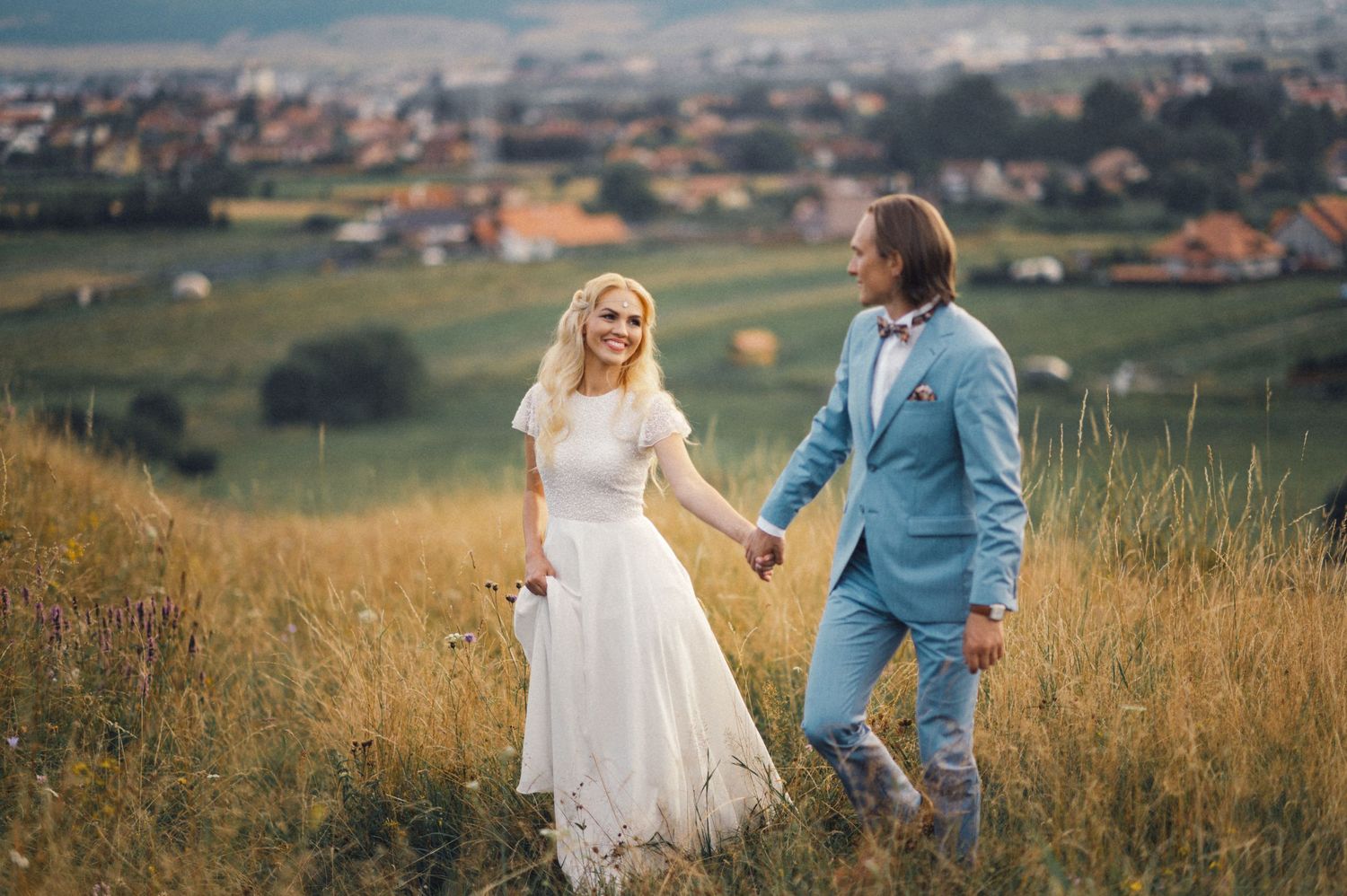 Newlyweds walking hand in hand through a hilltop meadow with a scenic town view in the background.