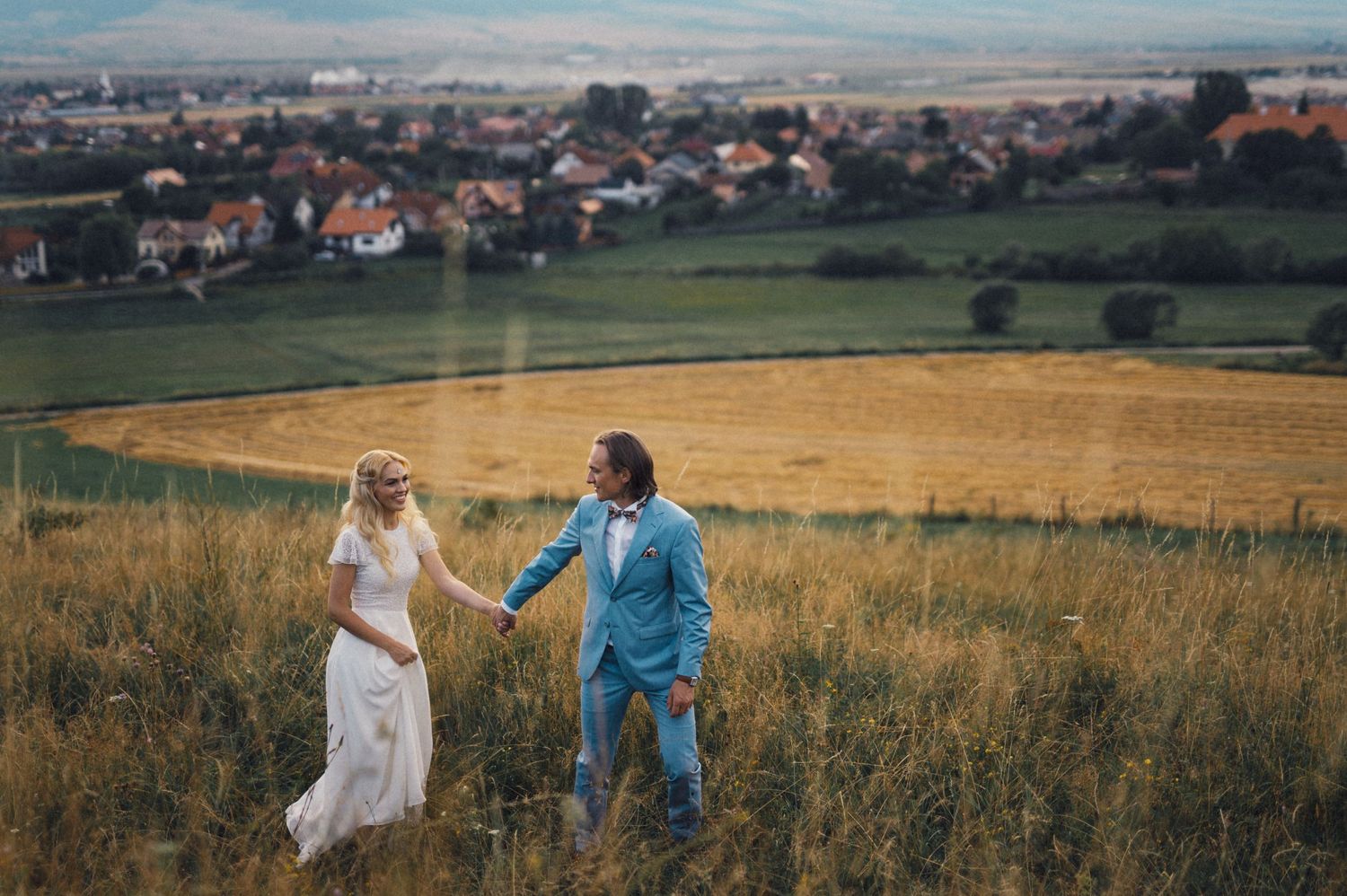 Wedding couple running playfully through a hillside meadow overlooking a scenic village landscape.