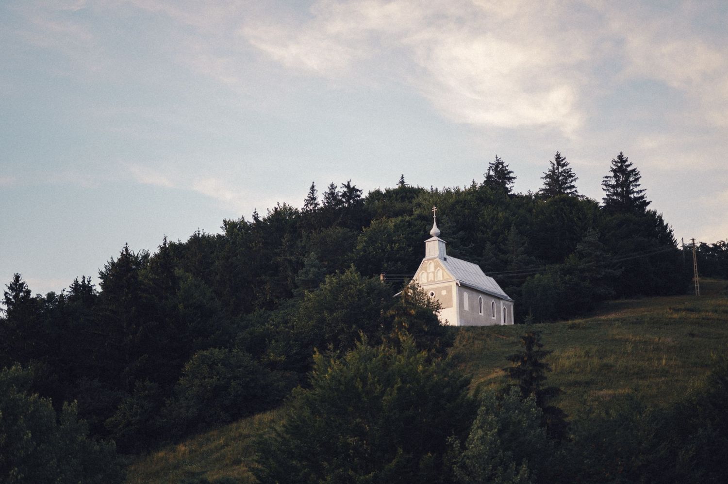 Small white chapel perched on a forested hillside under a cloudy sky.