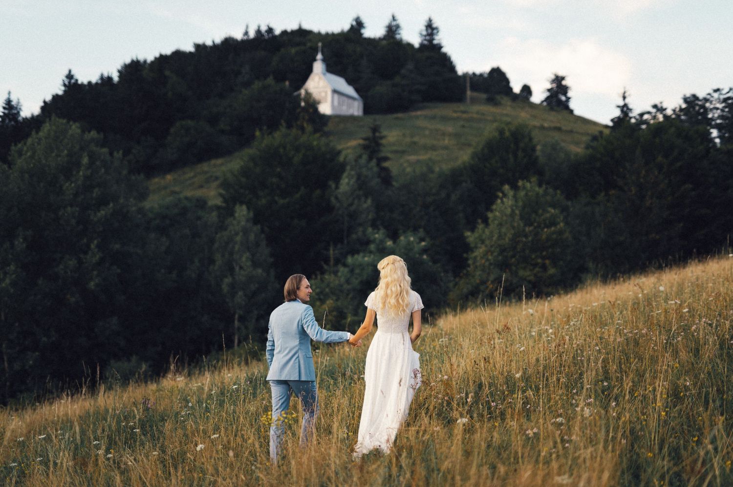 Couple walking up a grassy hill towards a white chapel nestled among pine trees.